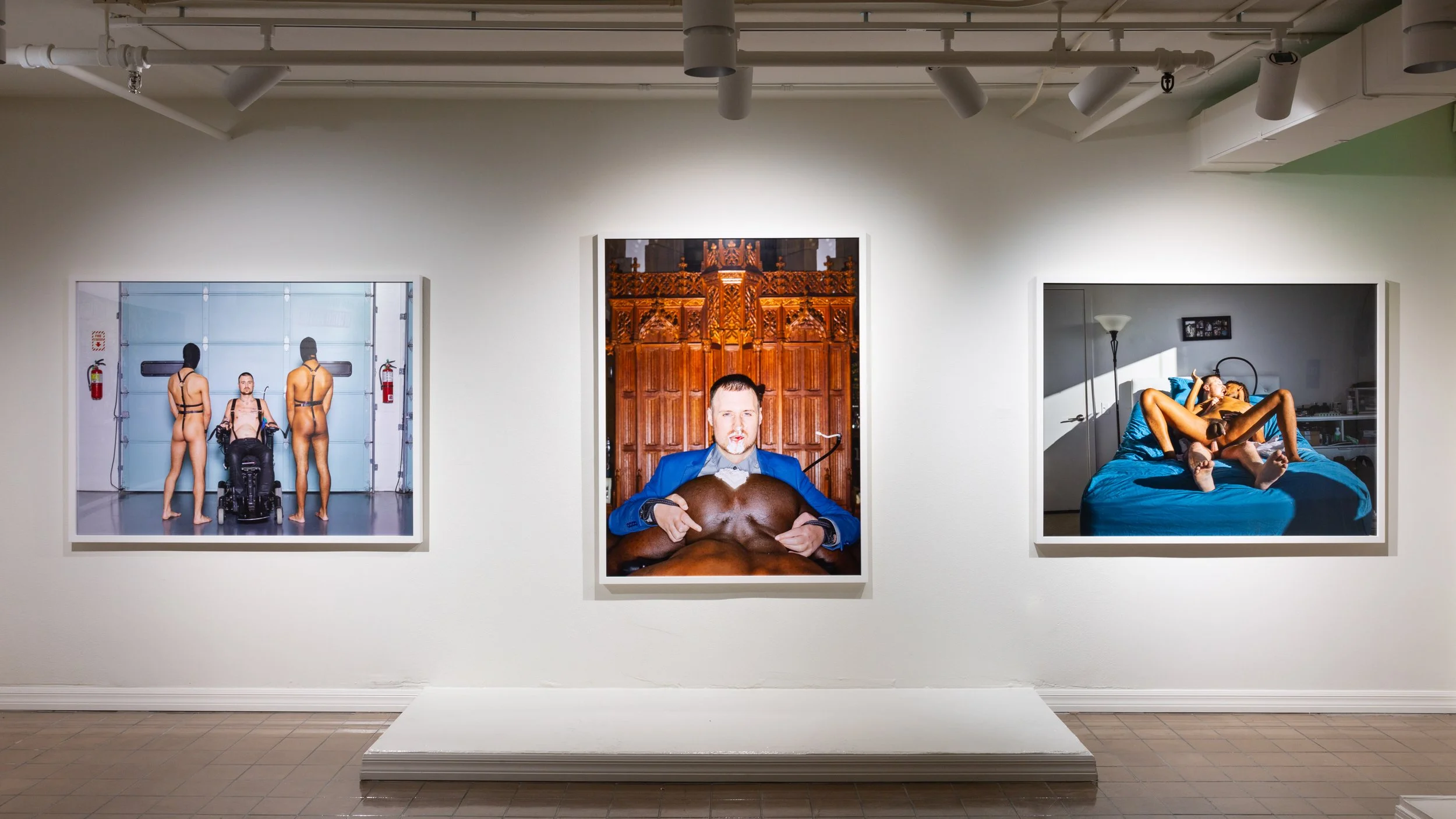 Three art photographs displayed on a gallery wall. The left photo shows three people in masks and minimal clothing in a garage. The middle photo depicts a man in a blue suit holding a large pig. The right photo features a woman lying on a bed with he