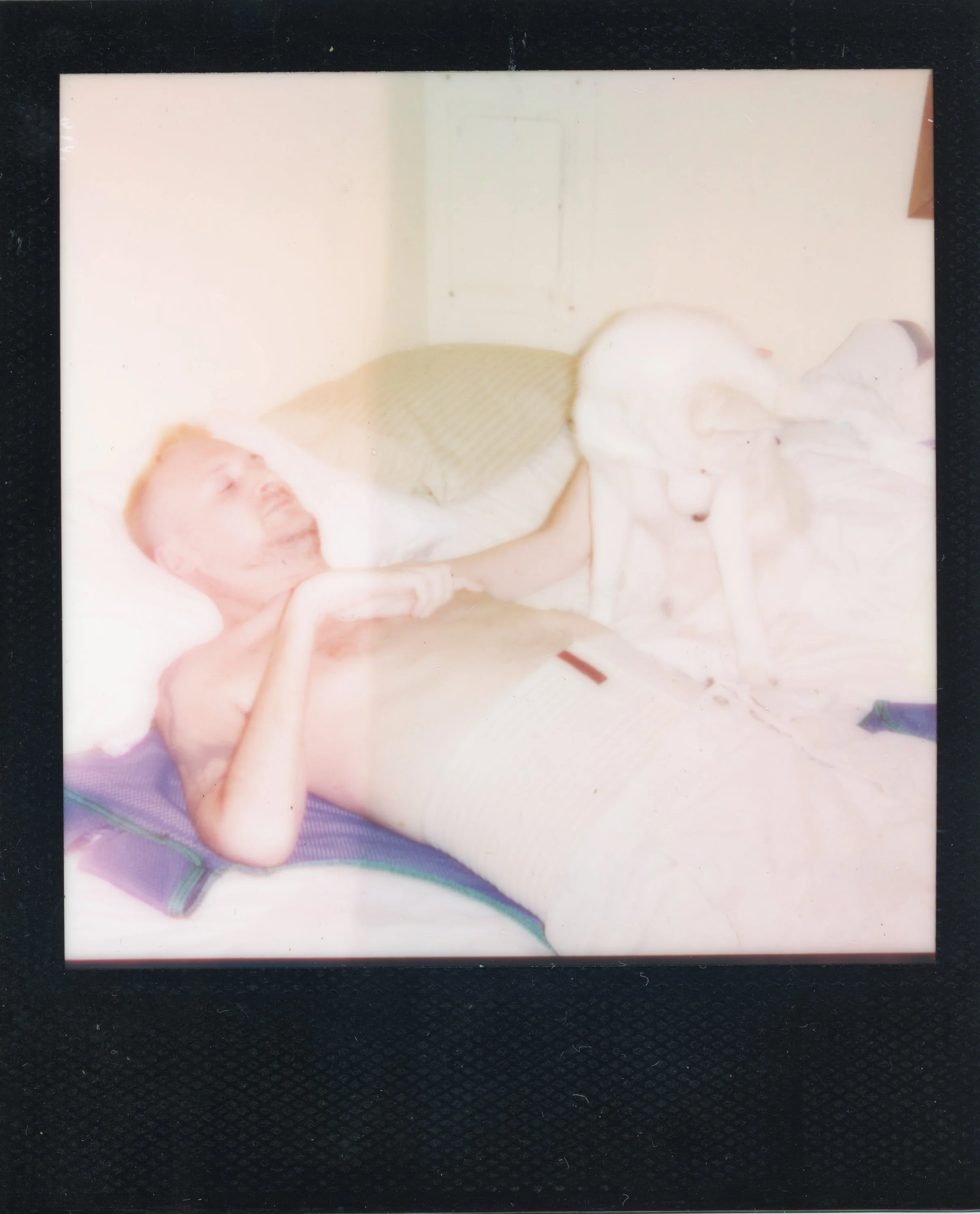 A distressed man lying shirtless on a bed with a blanket, holding a dog by the collar, with a woman bent over near his legs, in a room with a plain wall and a window
