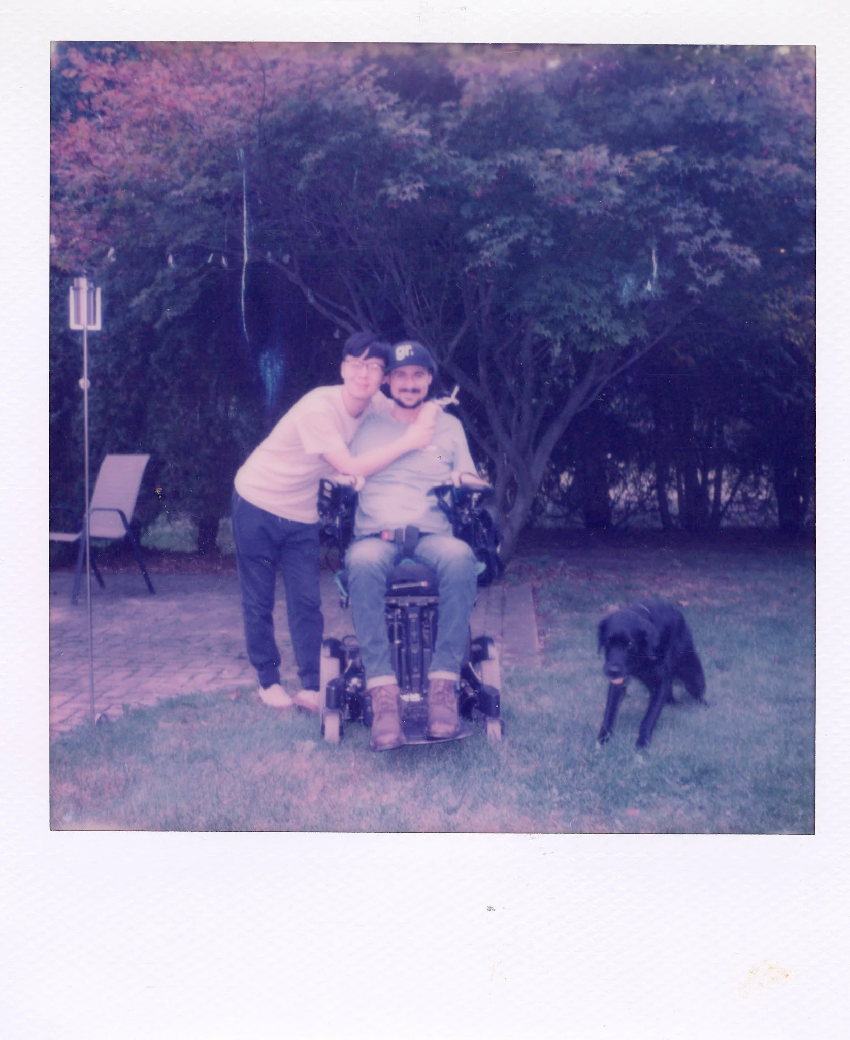 Two young men, one sitting in a wheelchair and the other standing beside him, smiling and hugging in a park with a large tree in the background. A black dog is walking on the grass nearby.