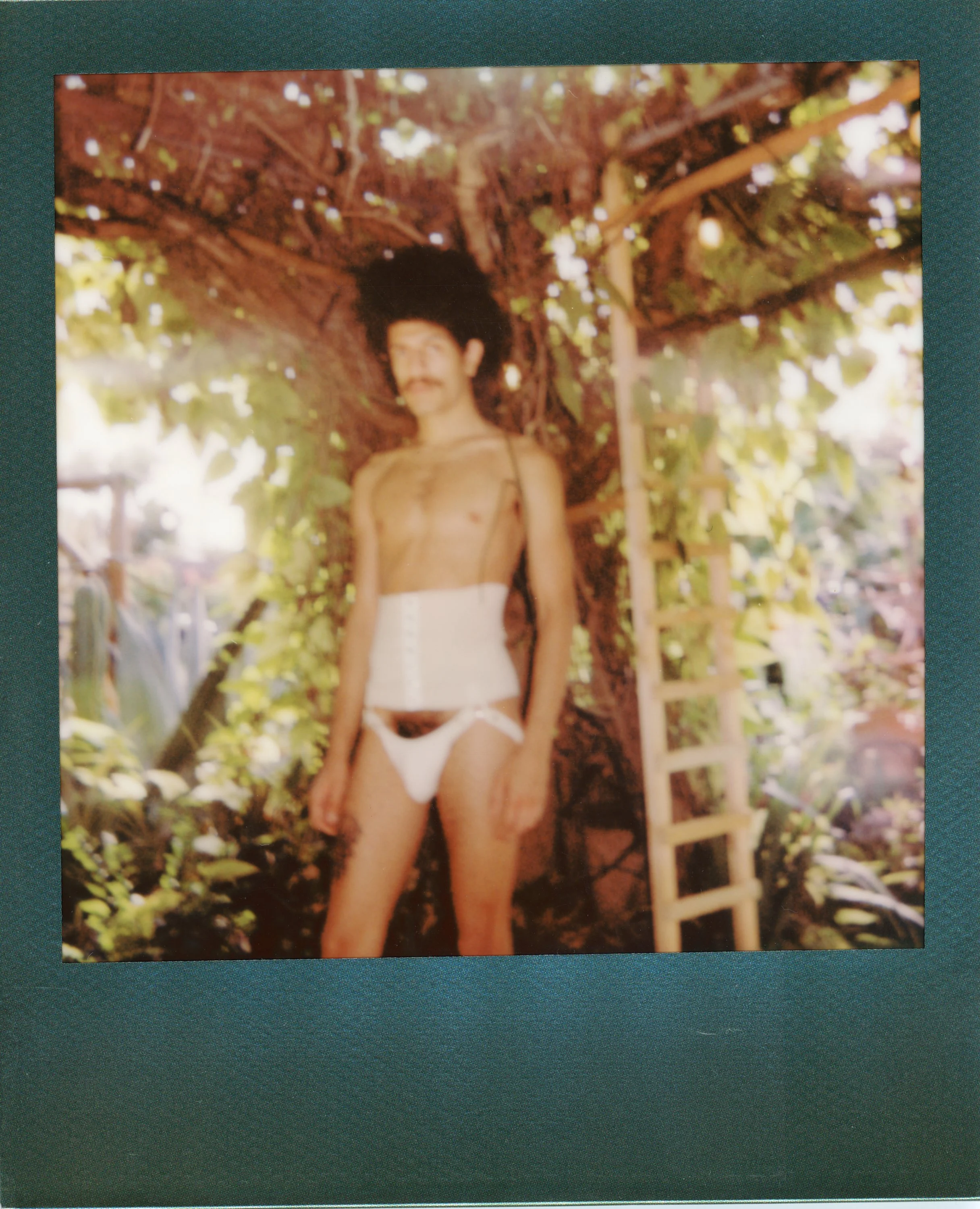 A man with a large black curly wig and mustache, shirtless, wearing a white corset and Petticoat, standing outdoors near a tree with green leaves and surrounded by plants.