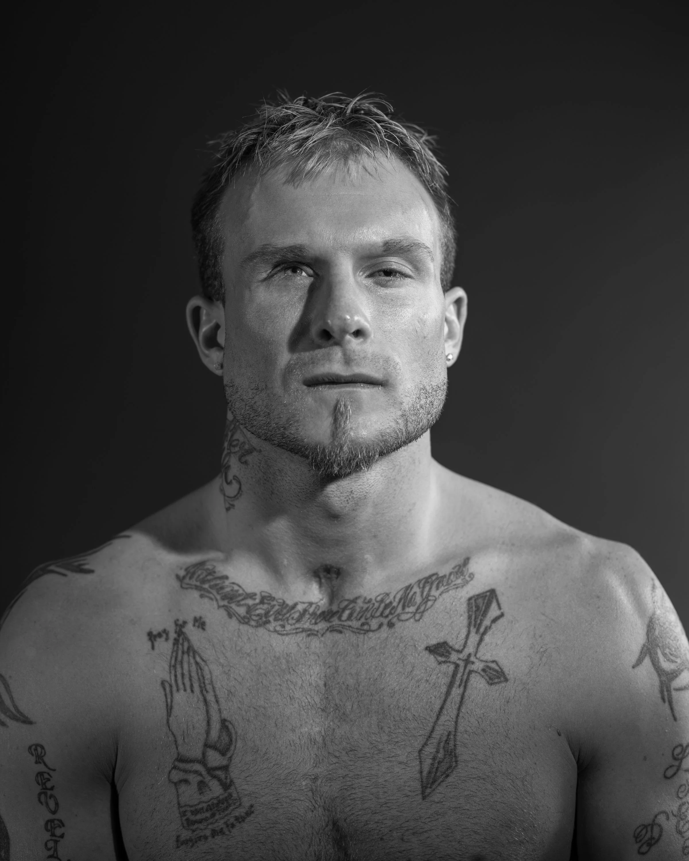 Black and white portrait of a shirtless man with tattoos, short spiky hair, and a goatee, looking directly at the camera with a serious expression.