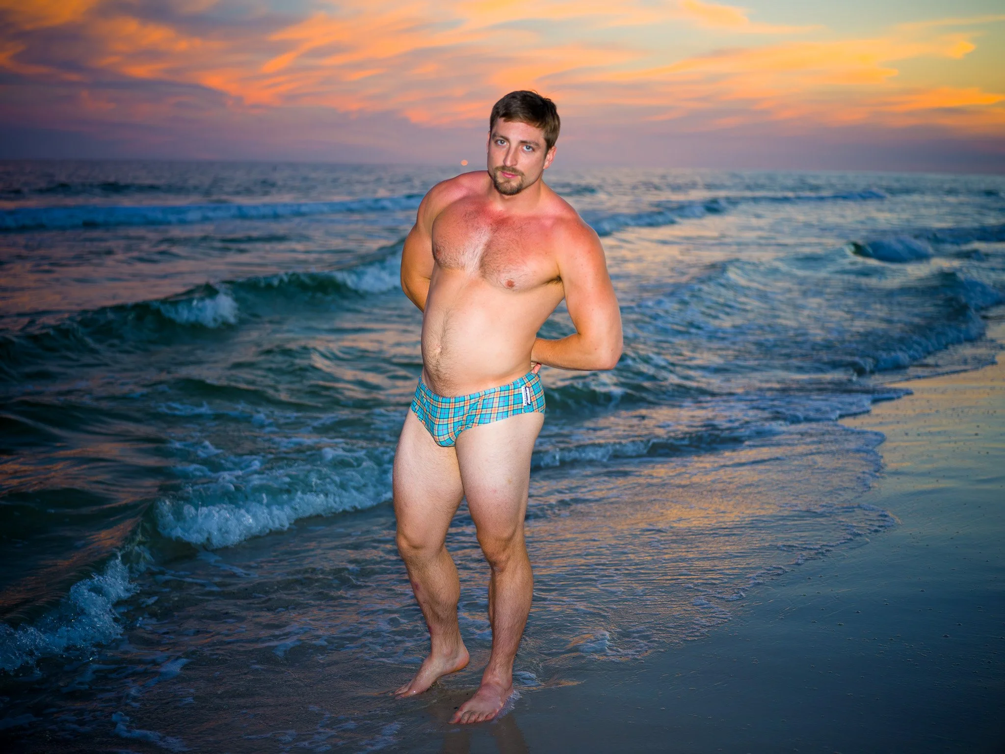 A man standing on a beach at sunset, wearing only plaid swimming trunks, with hands behind his back, looking at the camera.