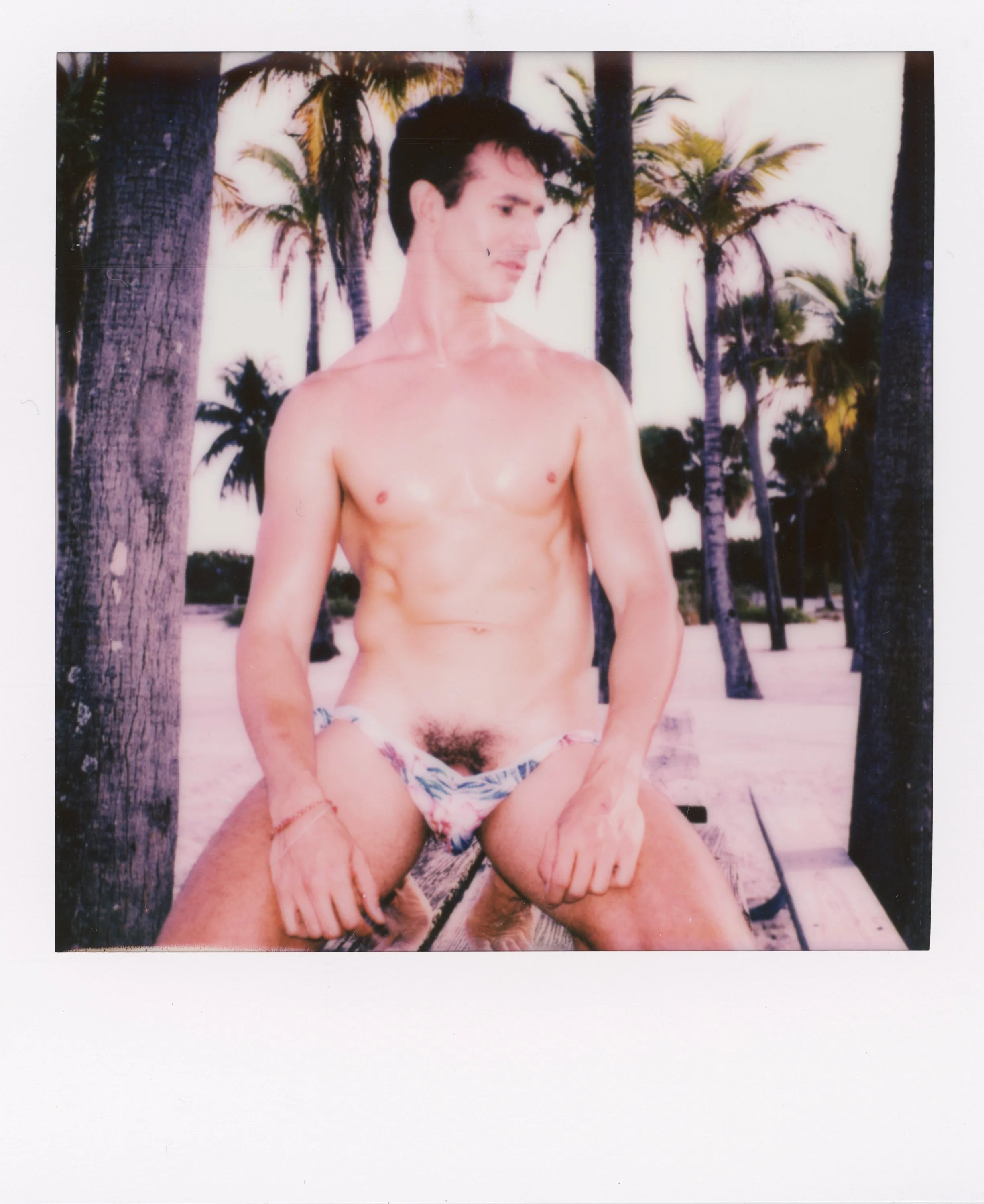 A young man is sitting on a wooden bench at the beach, wearing a floral bikini bottom and no shirt, with palm trees in the background.