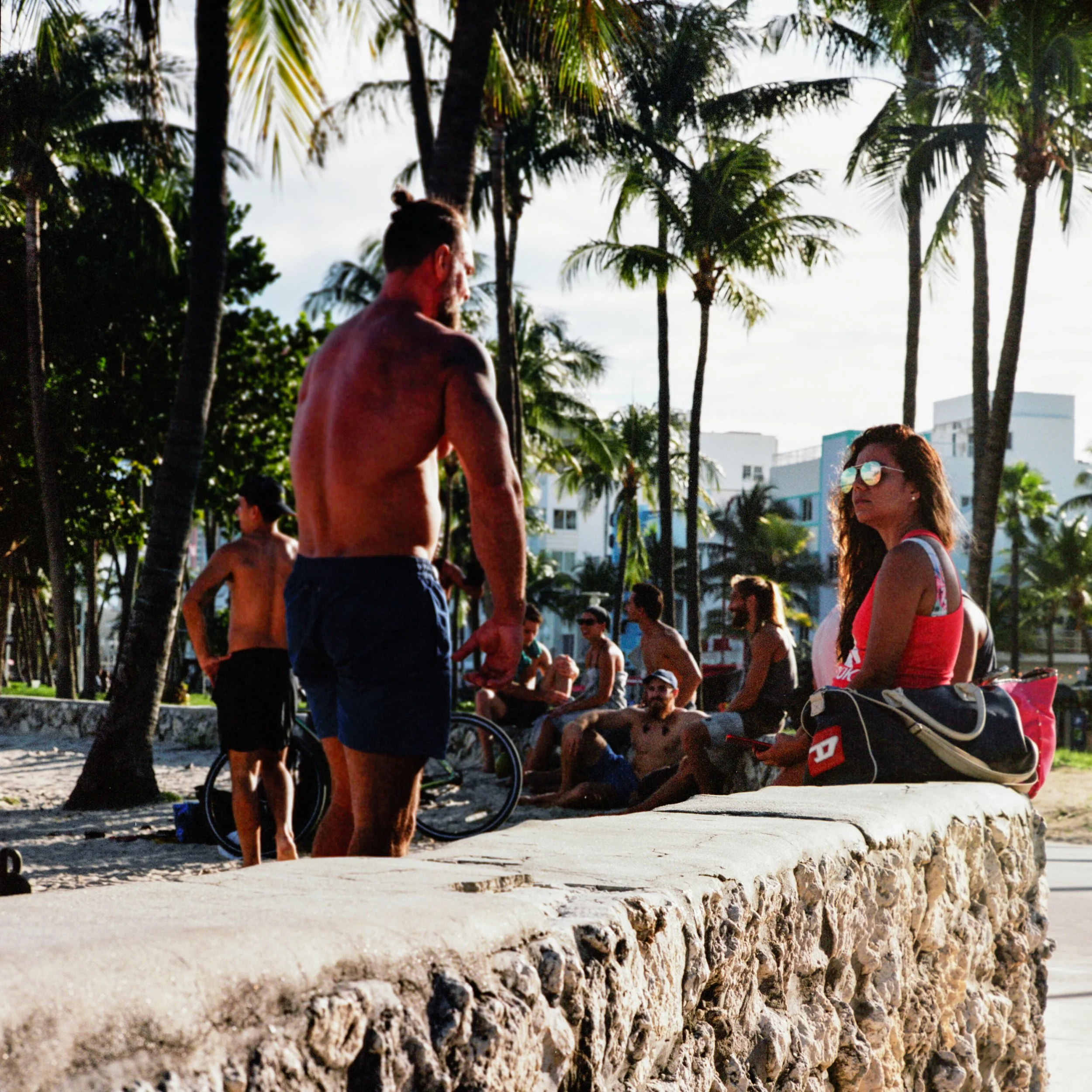 A man and woman sitting on a stone ledge in a park with palm trees, with other people sitting and walking nearby, suggesting a beach or coastal city environment.