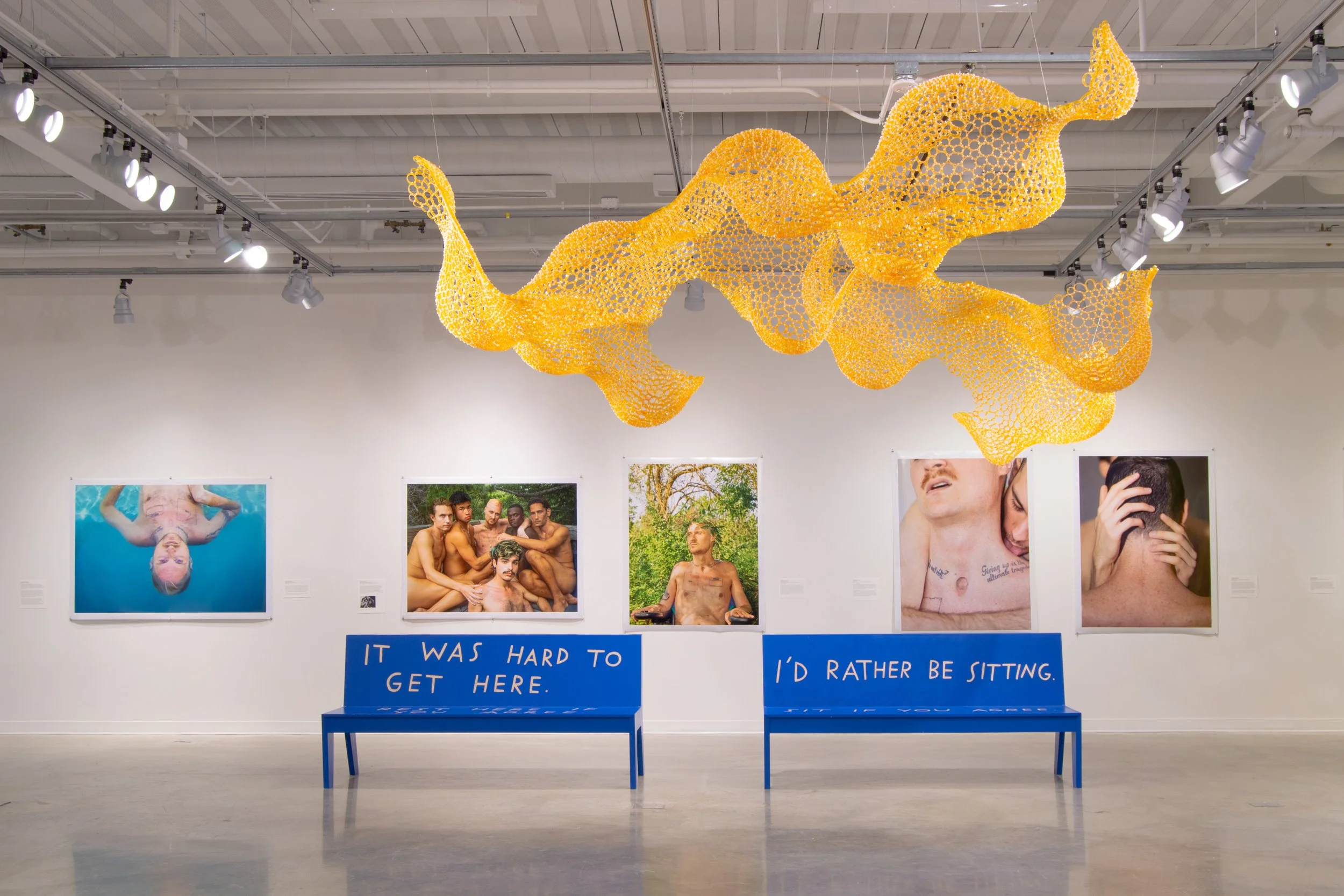 An art gallery display with five photographs of people, two blue benches with handwritten signs, and a large spiral-shaped yellow sculpture hanging from the ceiling.