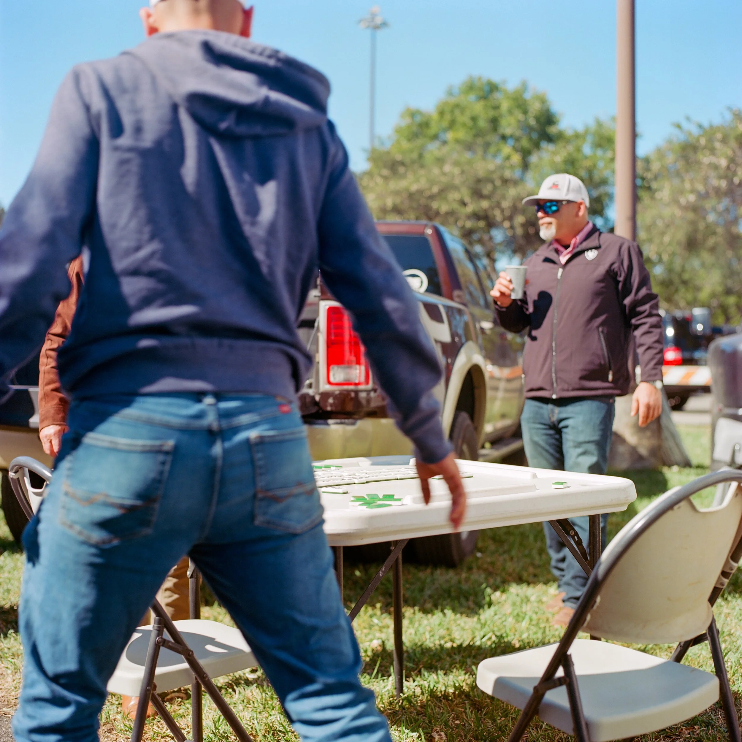 A person in a blue hoodie and jeans playing a game of giant Jenga at an outdoor event, with a man wearing sunglasses and a cap watching nearby.