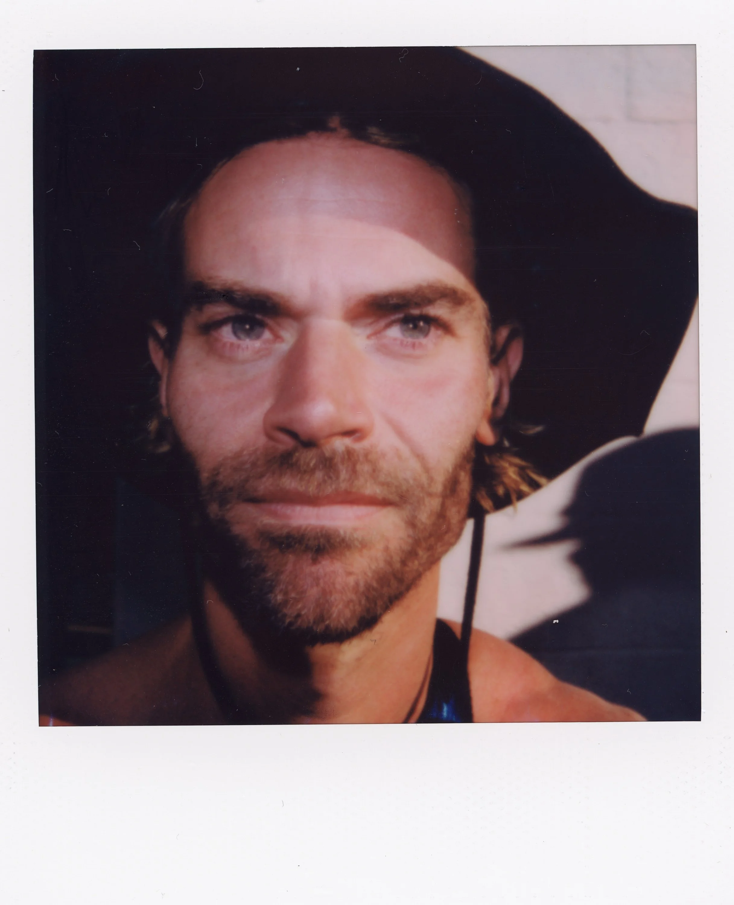 Close-up portrait of a man with blue eyes, light brown hair, a beard, wearing a black cowboy hat and a necklace, smiling slightly.