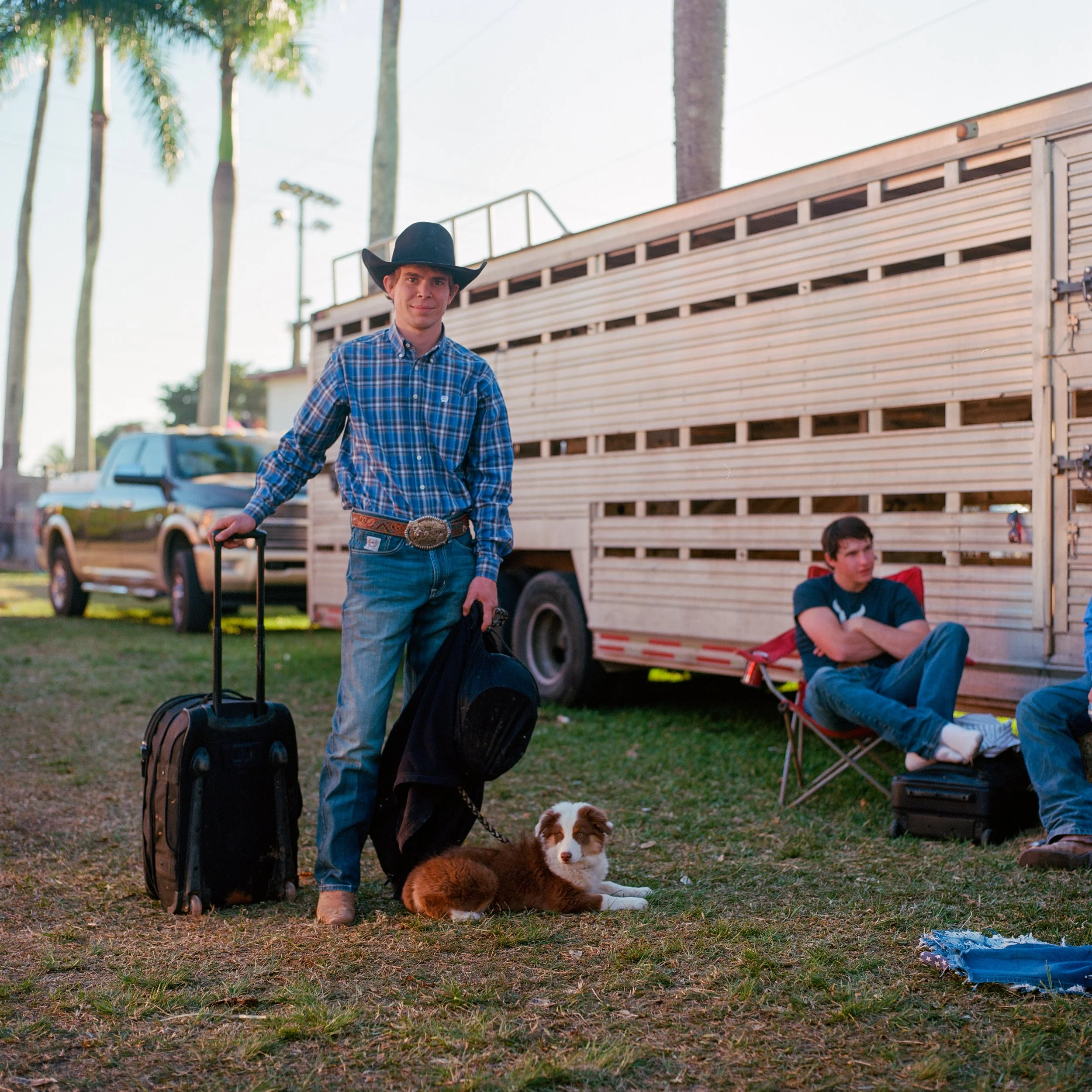 Young man in cowboy attire with a hat, plaid shirt, and jeans standing on grass with a suitcase and a dog, with a large trailer and another person sitting in a foldable chair in the background.