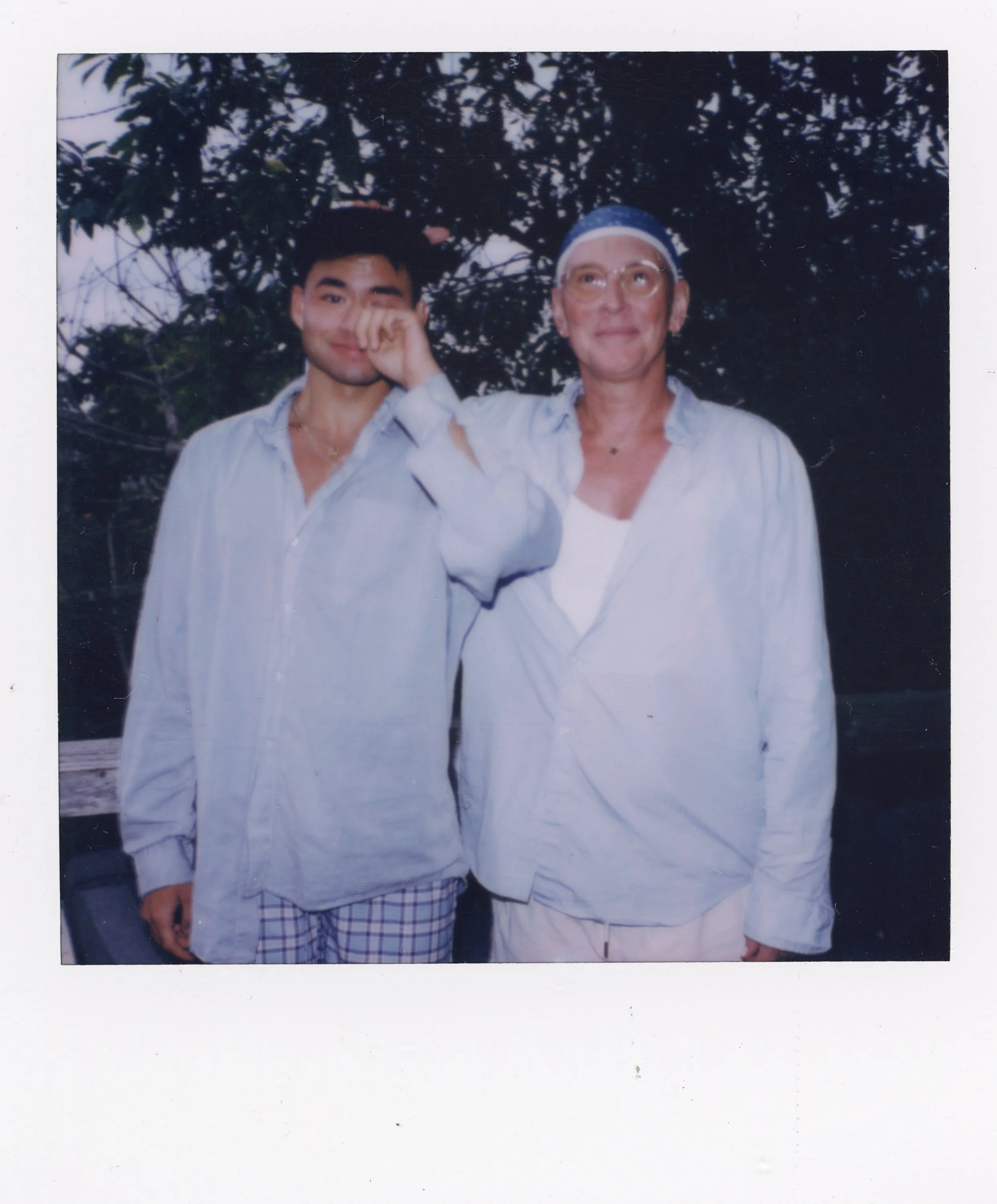 A young man and an older woman standing outdoors in front of trees, with the young man touching the woman's head and both smiling, dressed in light-colored casual clothing.