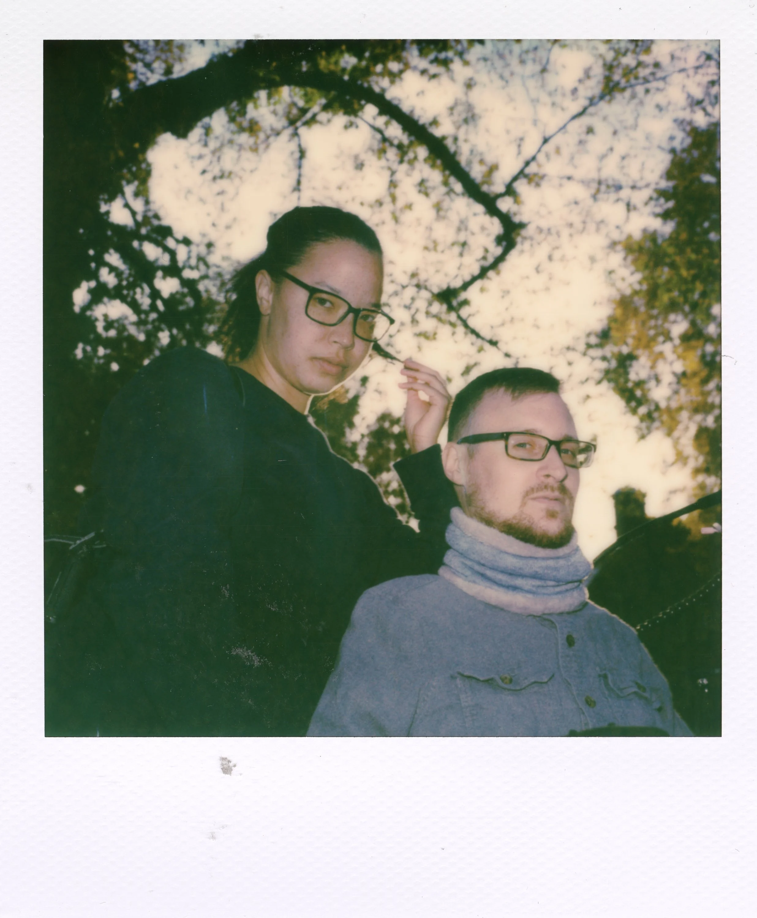 Two people, a woman and a man, wearing glasses, outdoors with trees and sky in the background.