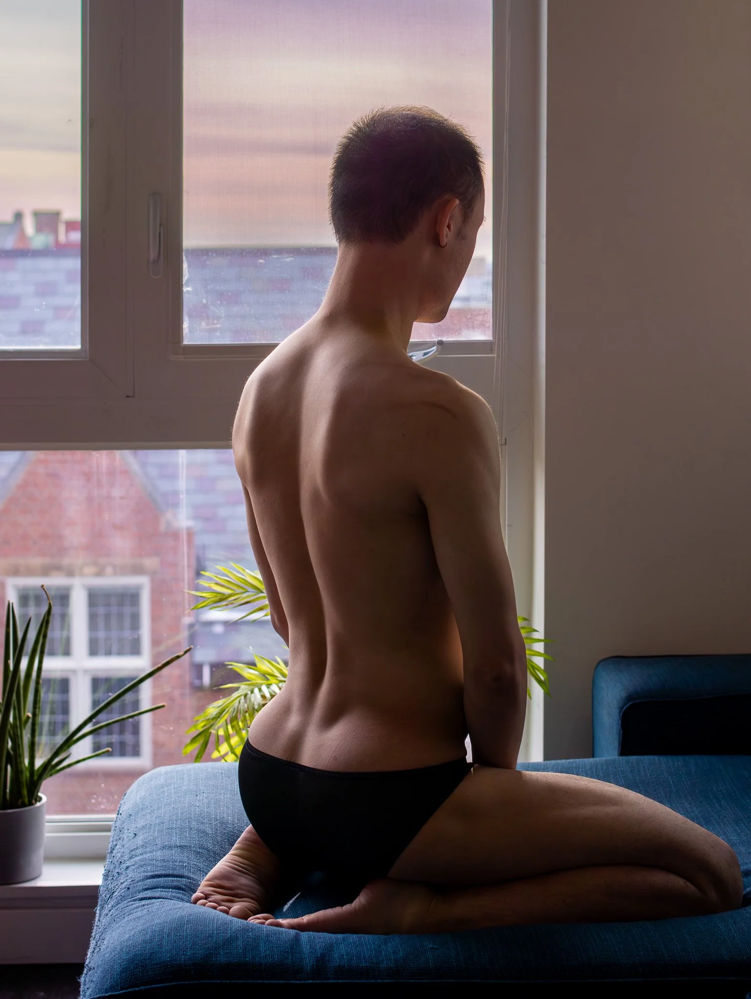 A person with short dark hair kneeling on a blue sofa, facing a window with their back to the camera, wearing black underwear, and looking outside at a cityscape with buildings and a pastel sky.