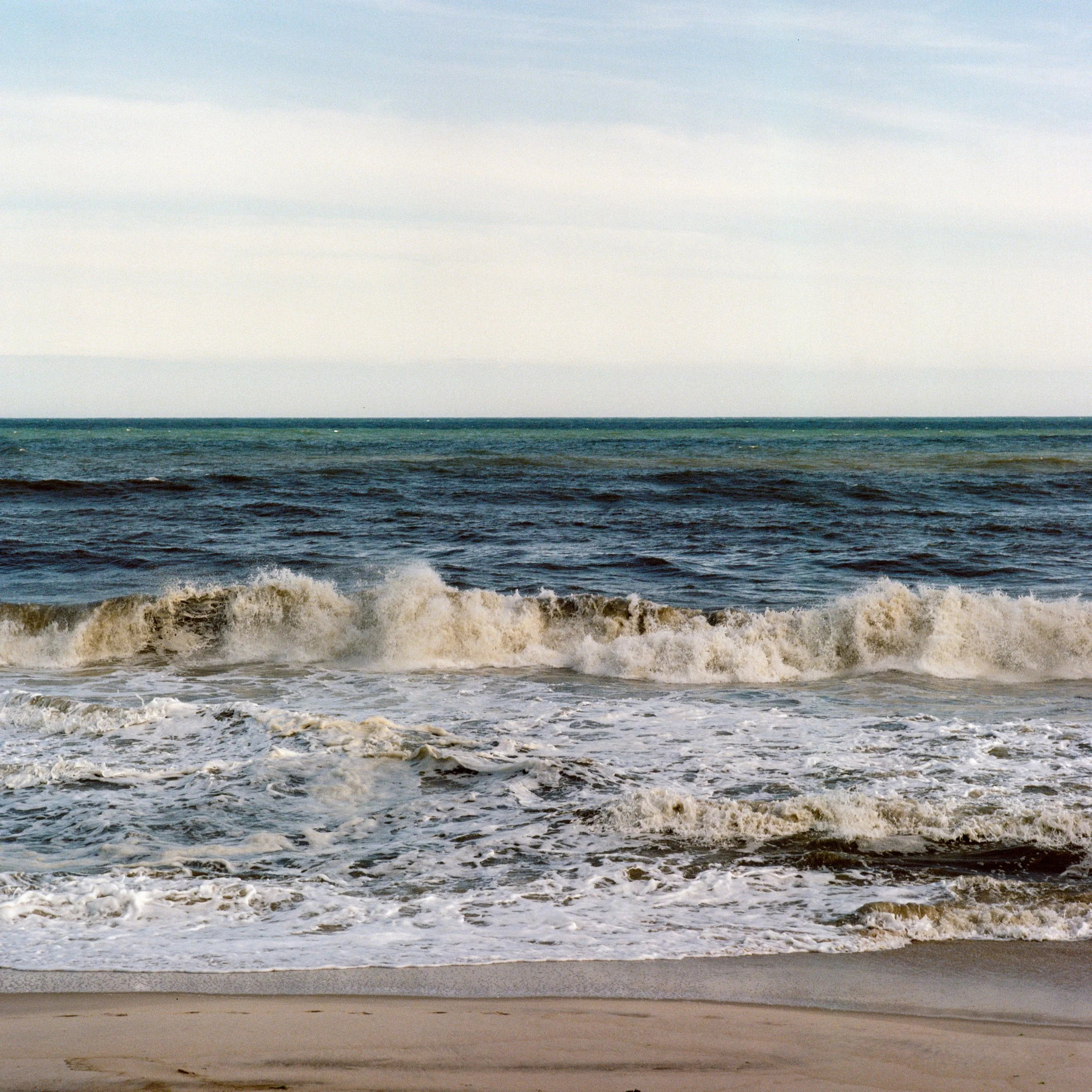 Waves crashing on the sandy shore of a beach with the ocean extending to the horizon under a partly cloudy sky.