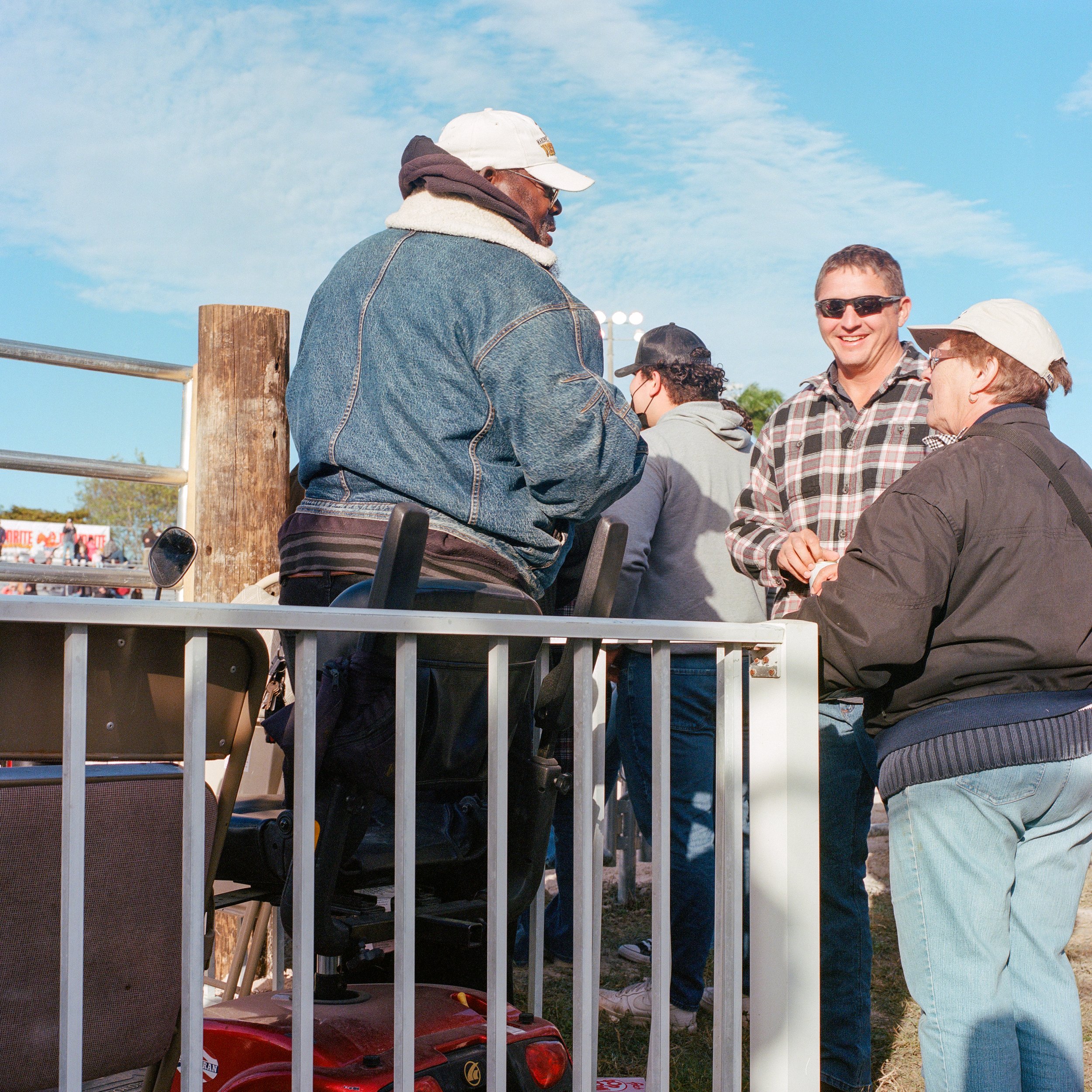 Group of people outdoors, talking and laughing near a wooden fence, under a blue sky