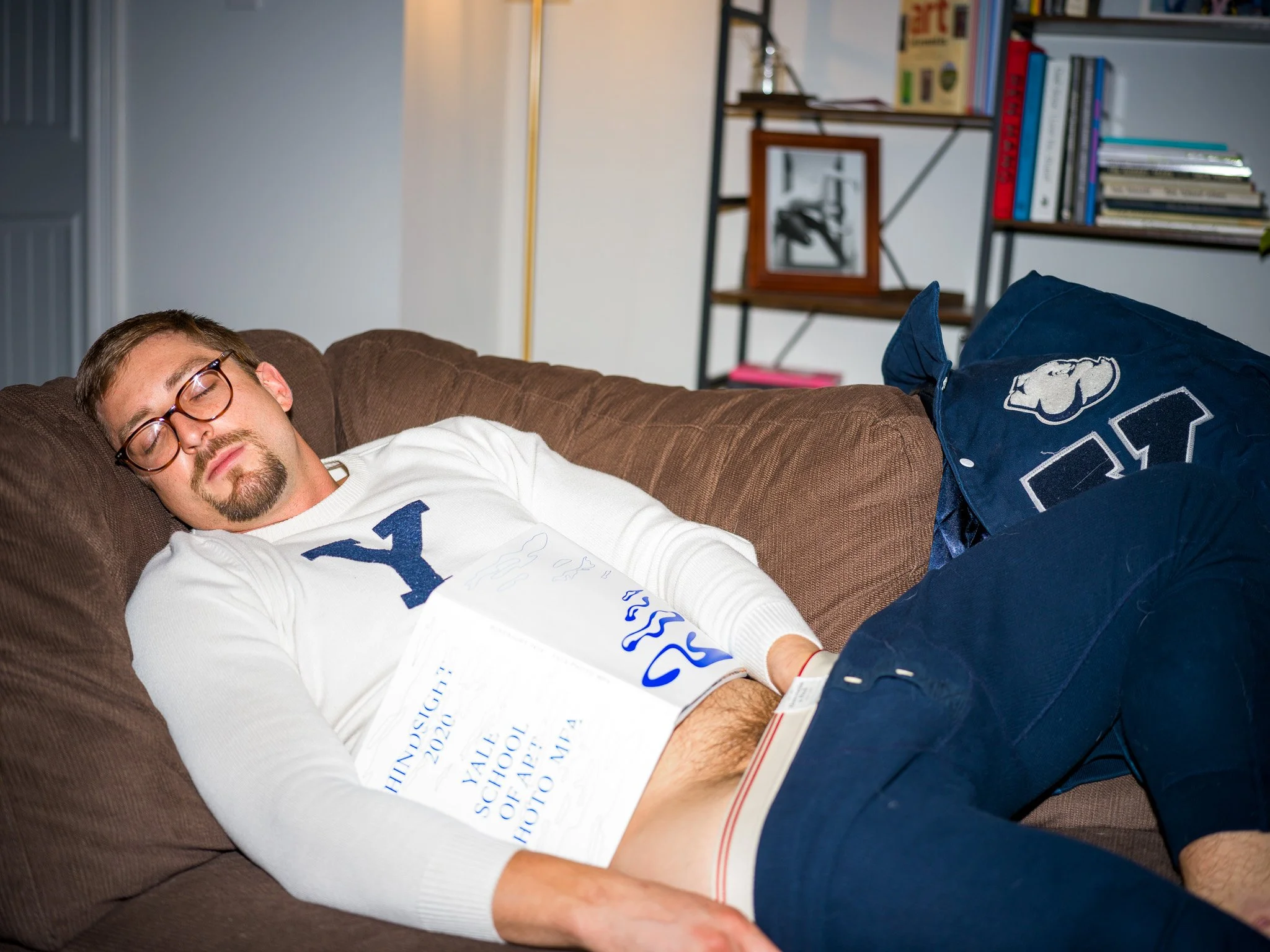 A man with glasses sleeping on a brown couch, holding a university diploma. He is dressed in a white sweater with a large blue letter 'Y' and dark pants, with a university bag on the couch beside him. In the background, there are bookshelves and fram