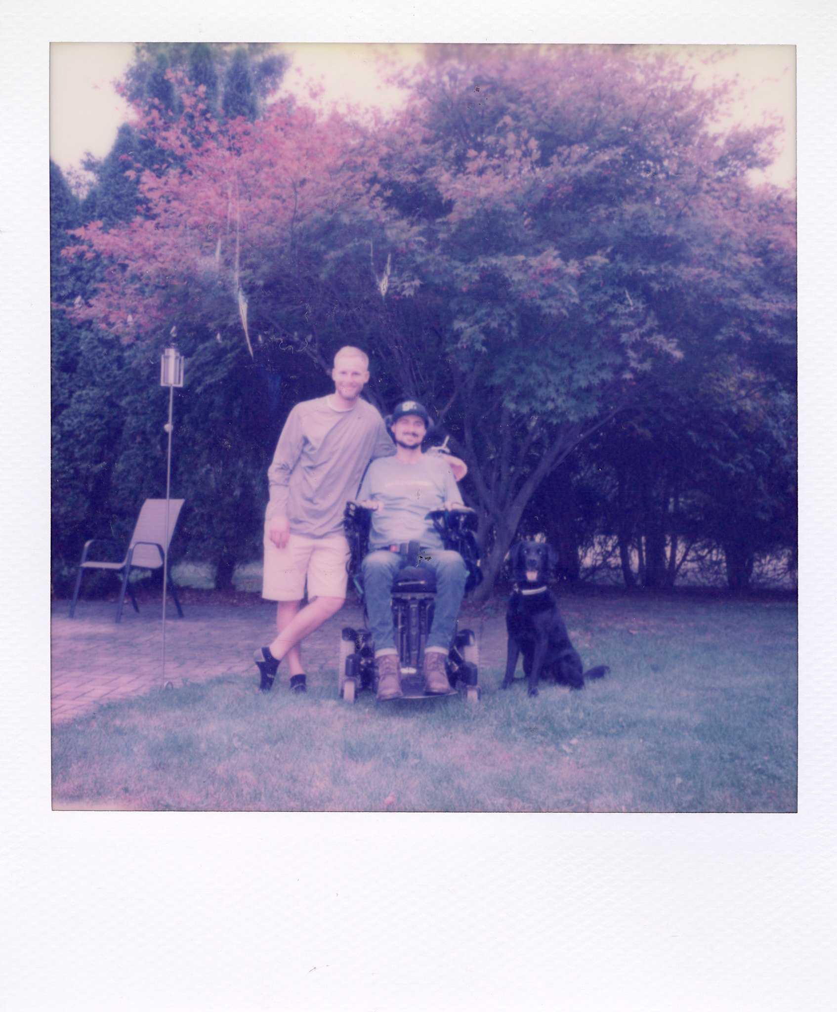 Two men and a dog outdoors in front of trees, one man in a wheelchair and one standing, with a chair and a bird feeder in the background.