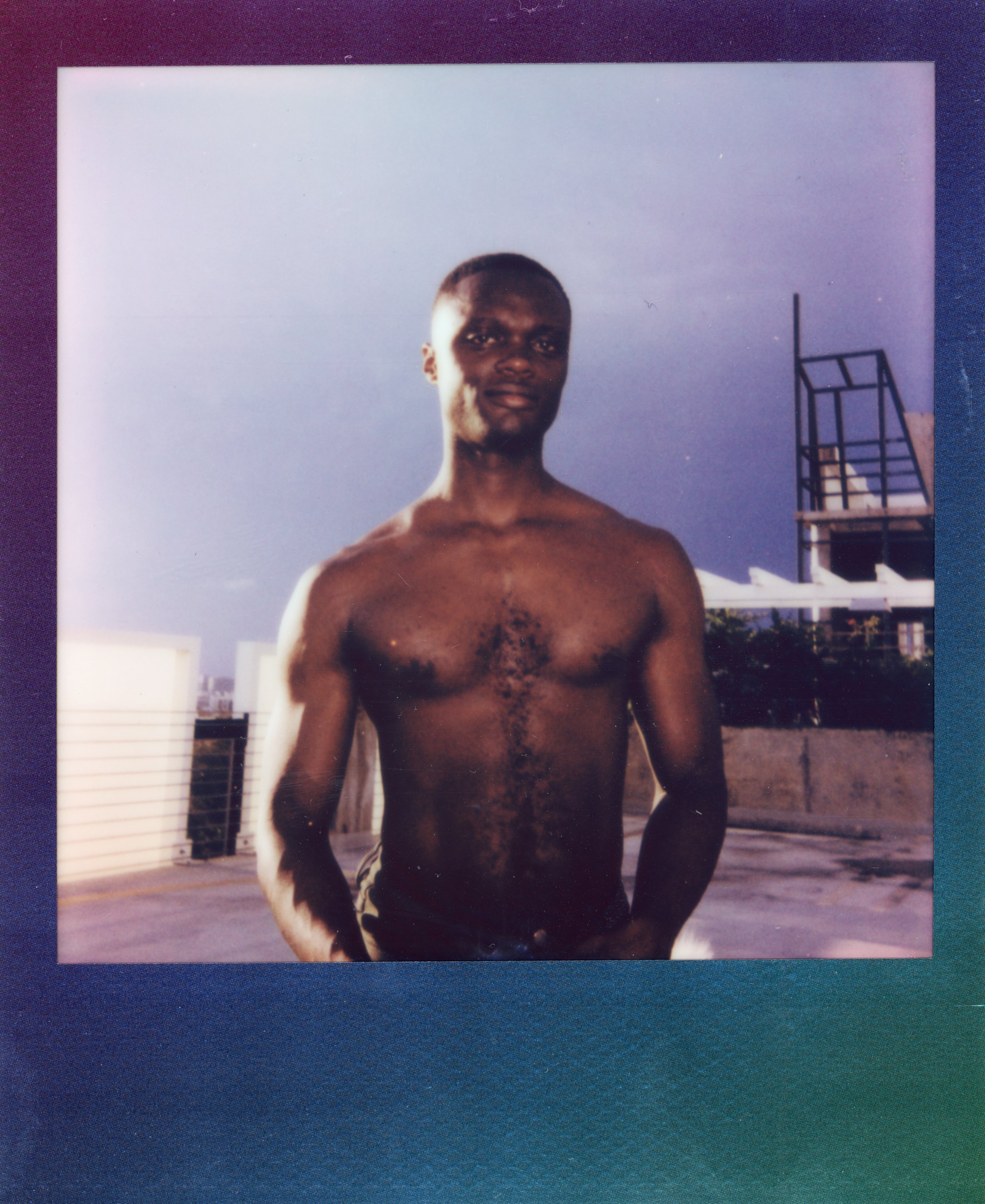 A shirtless young man standing outdoors on a rooftop with a blue sky background.