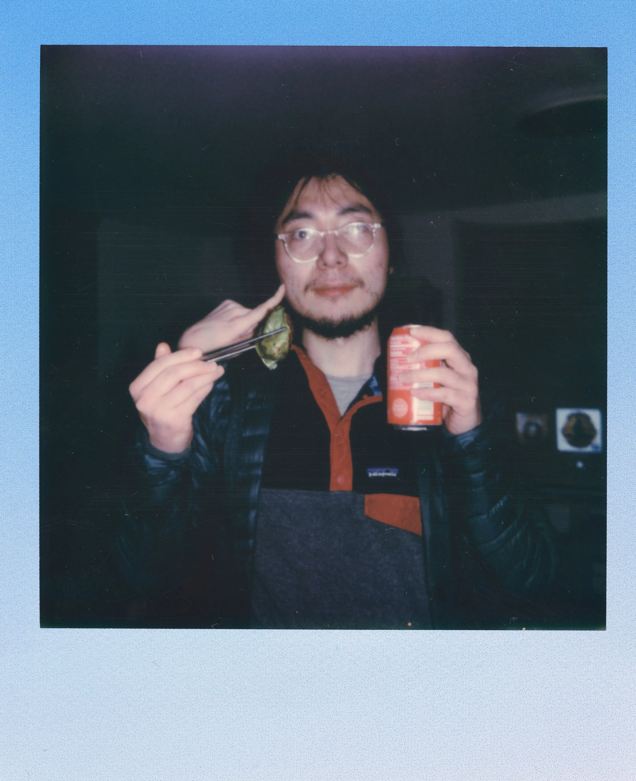 A man wearing glasses and a black jacket holds a can of soda in one hand and a piece of food with chopsticks in the other, standing indoors.