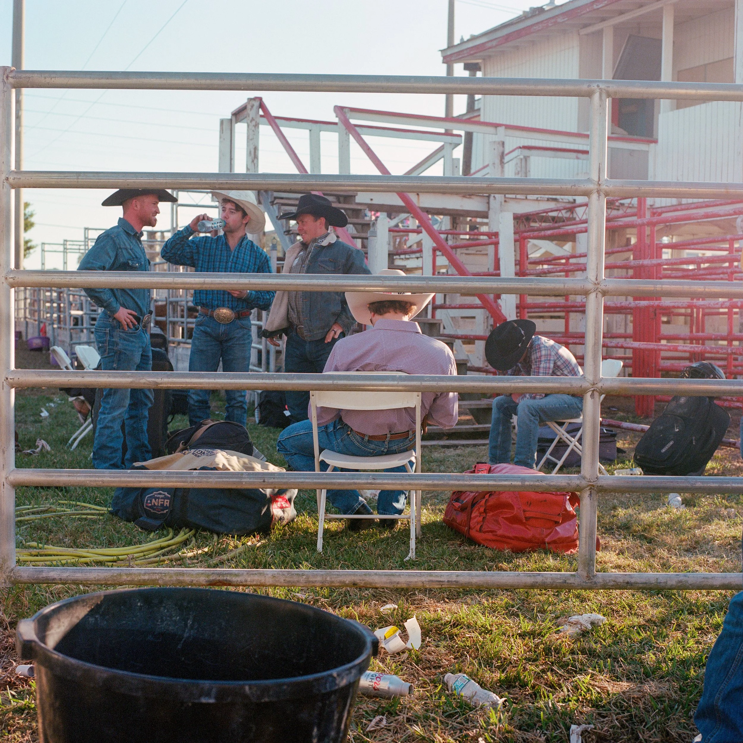 Five men wearing cowboy hats and jeans are gathered outside near a metal fence. Four men are standing and one is seated, drinking, talking and working, with some gear and bags on the ground. The scene is set outdoors near a barn or stable with a wood