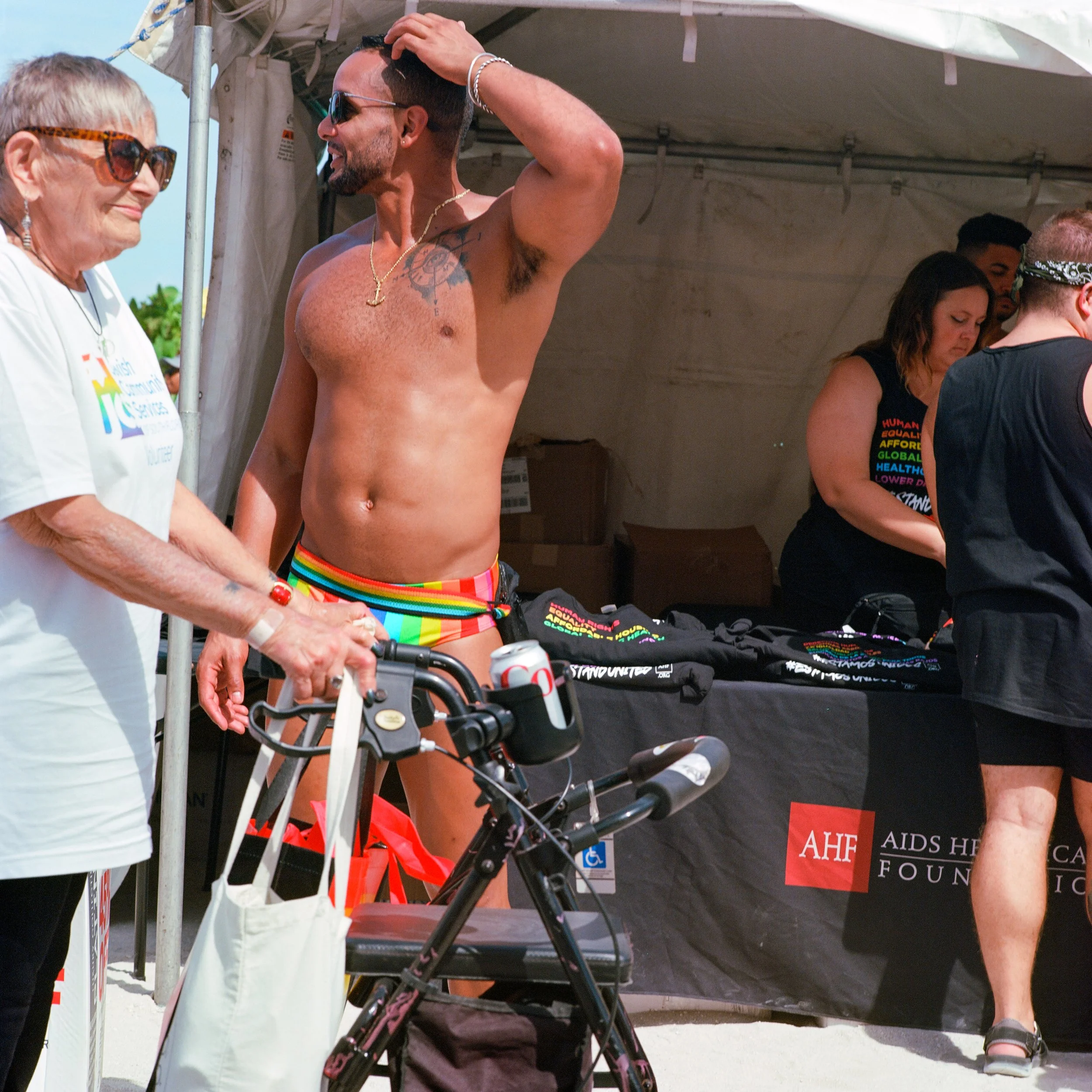 A man with sunglasses, tattoos, and rainbow swim trunks standing under a tent at an AIDS foundation event, talking to an elderly woman with glasses and a white T-shirt, next to a bicycle and a table with AIDS awareness merchandise.