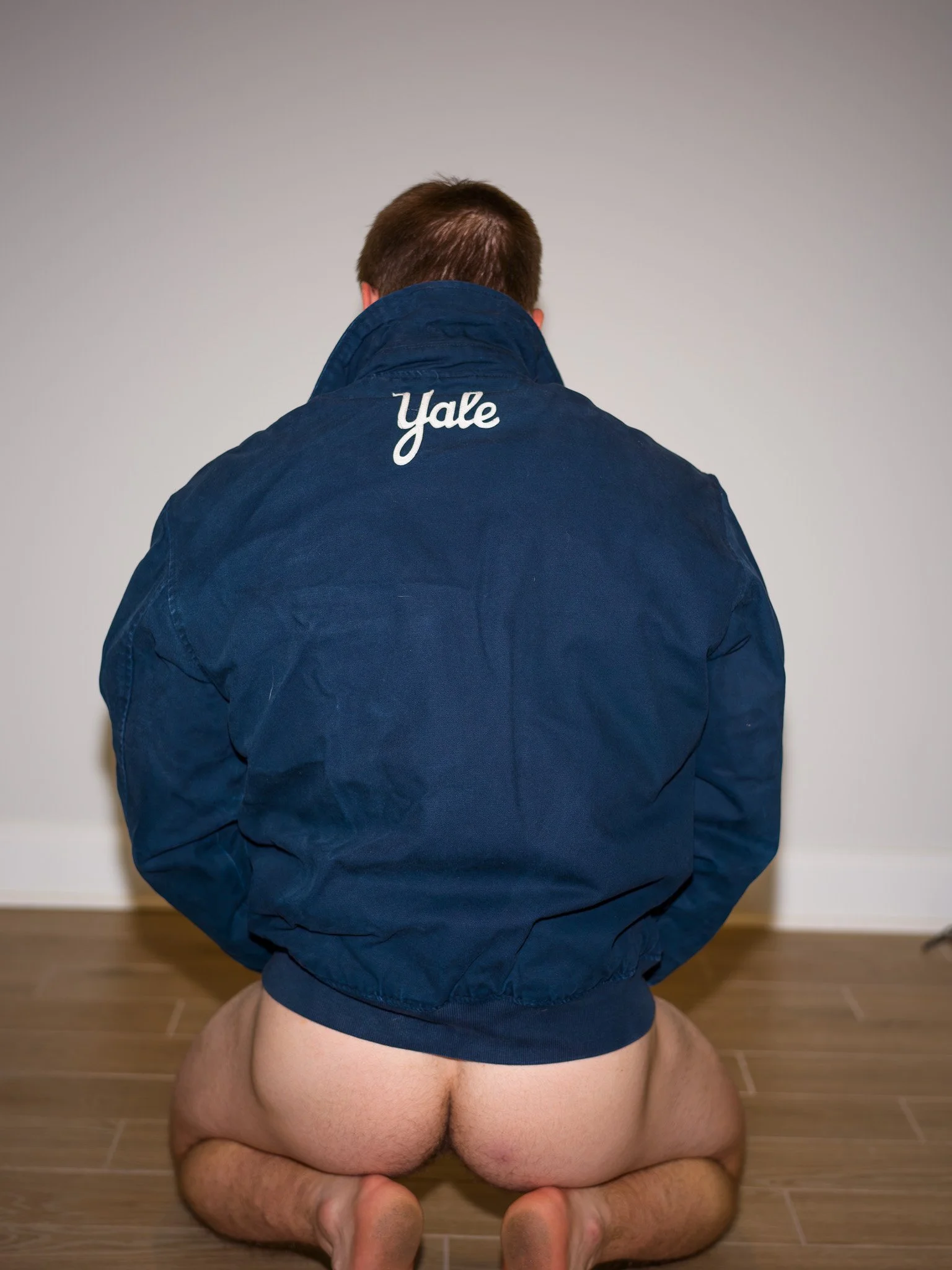 A person kneeling on the floor with their back facing the camera, wearing a blue Yale jacket, with their pants pulled down revealing their buttocks.