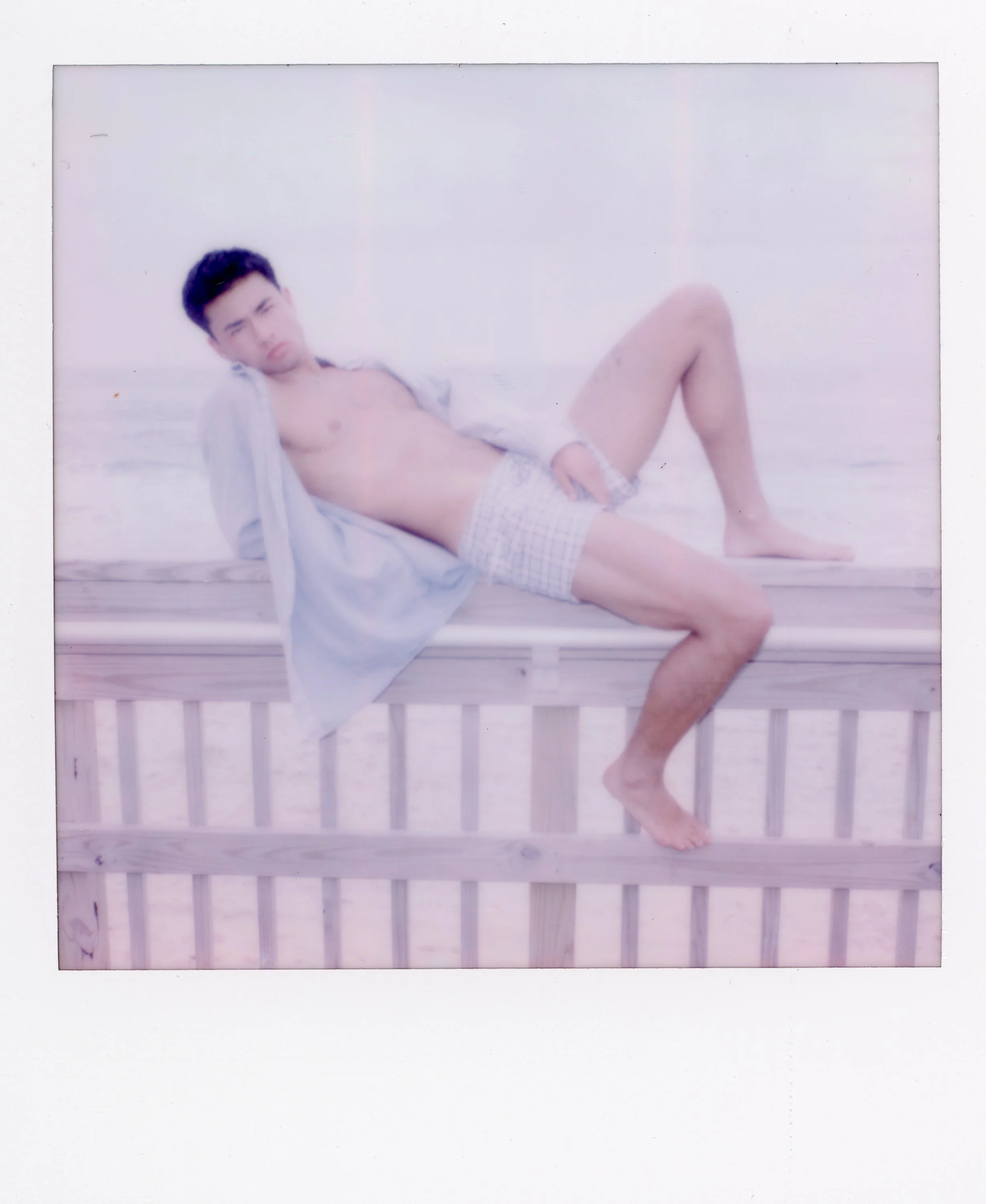 A young man reclining on a wooden railing at the beach, wearing an open shirt and plaid boxer shorts, with a relaxed pose and a slightly serious expression.