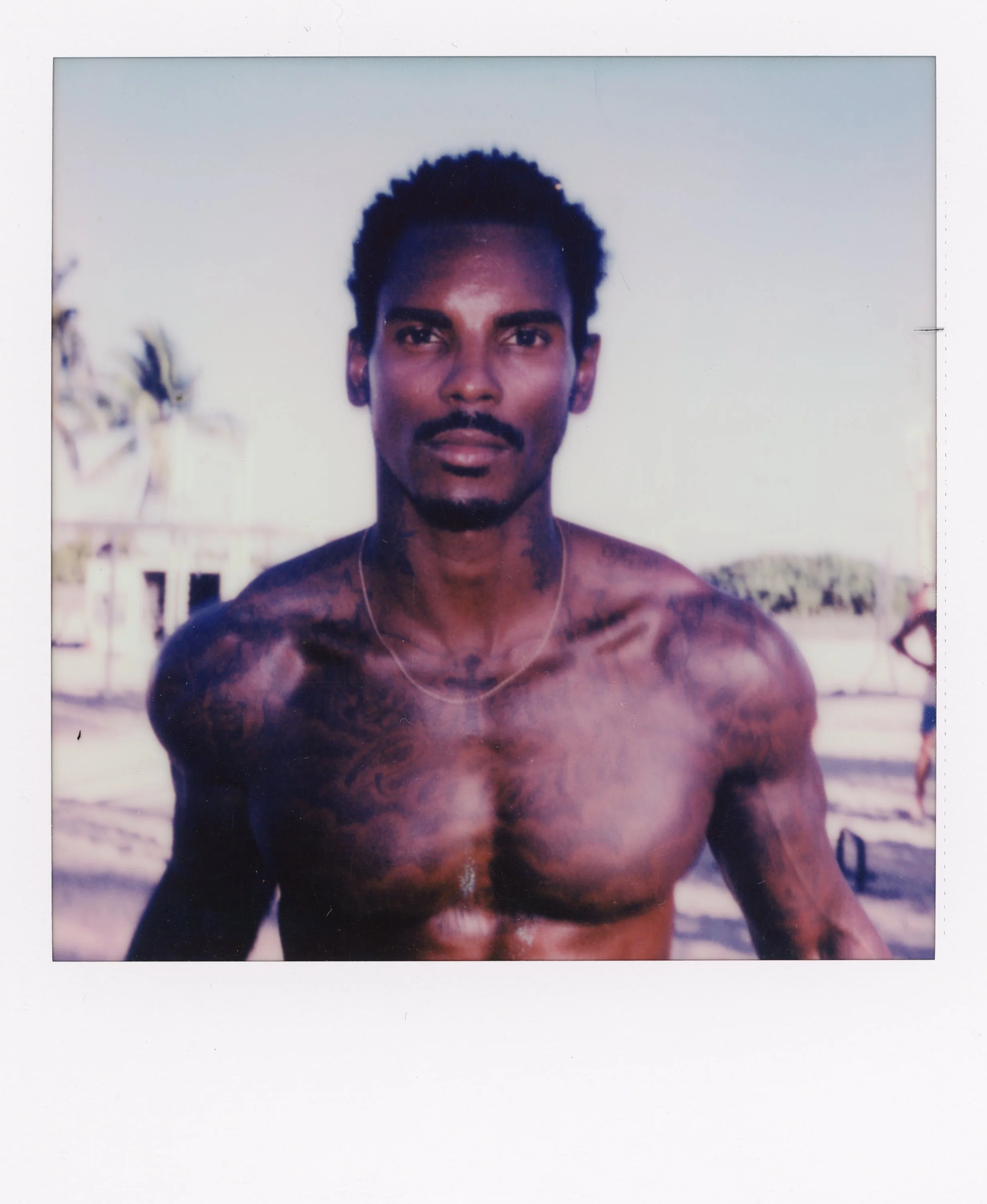 A shirtless man with tattoos on his shoulders and chest, standing outdoors on a beach with palm trees in the background.
