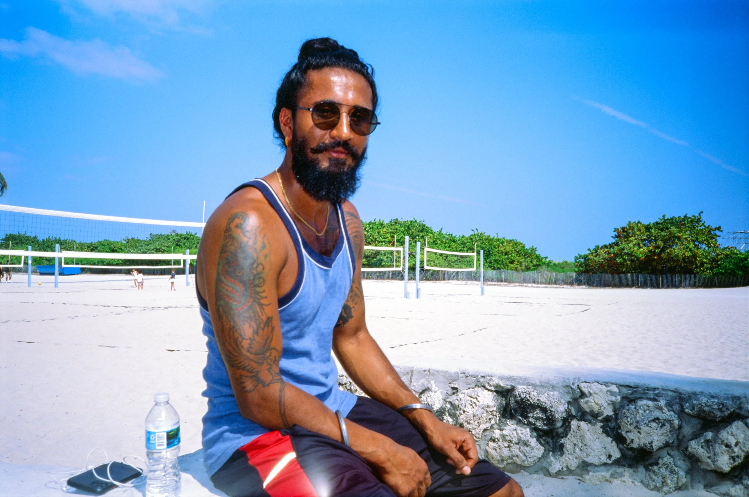 A man with tattoos, beard, and sunglasses sitting on a stone wall at a beach volleyball court under a blue sky with green trees in the background, sunscreen bottle, and face mask on the ground near him.