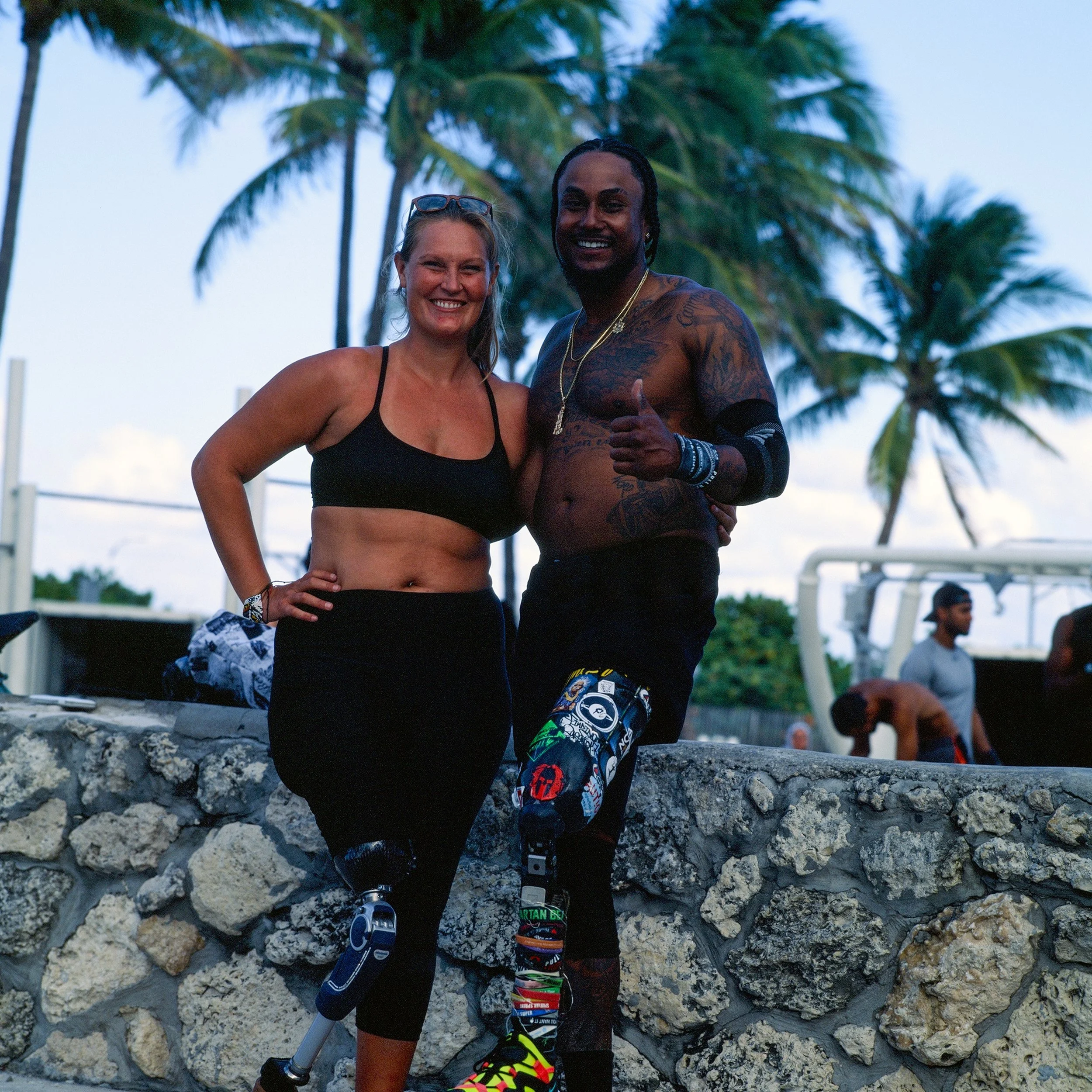 A smiling woman in a black sports bra and leggings with a prosthetic leg, standing next to a smiling shirtless man with tattoos and a prosthetic leg, giving a thumbs up, with palm trees and other people in the background.