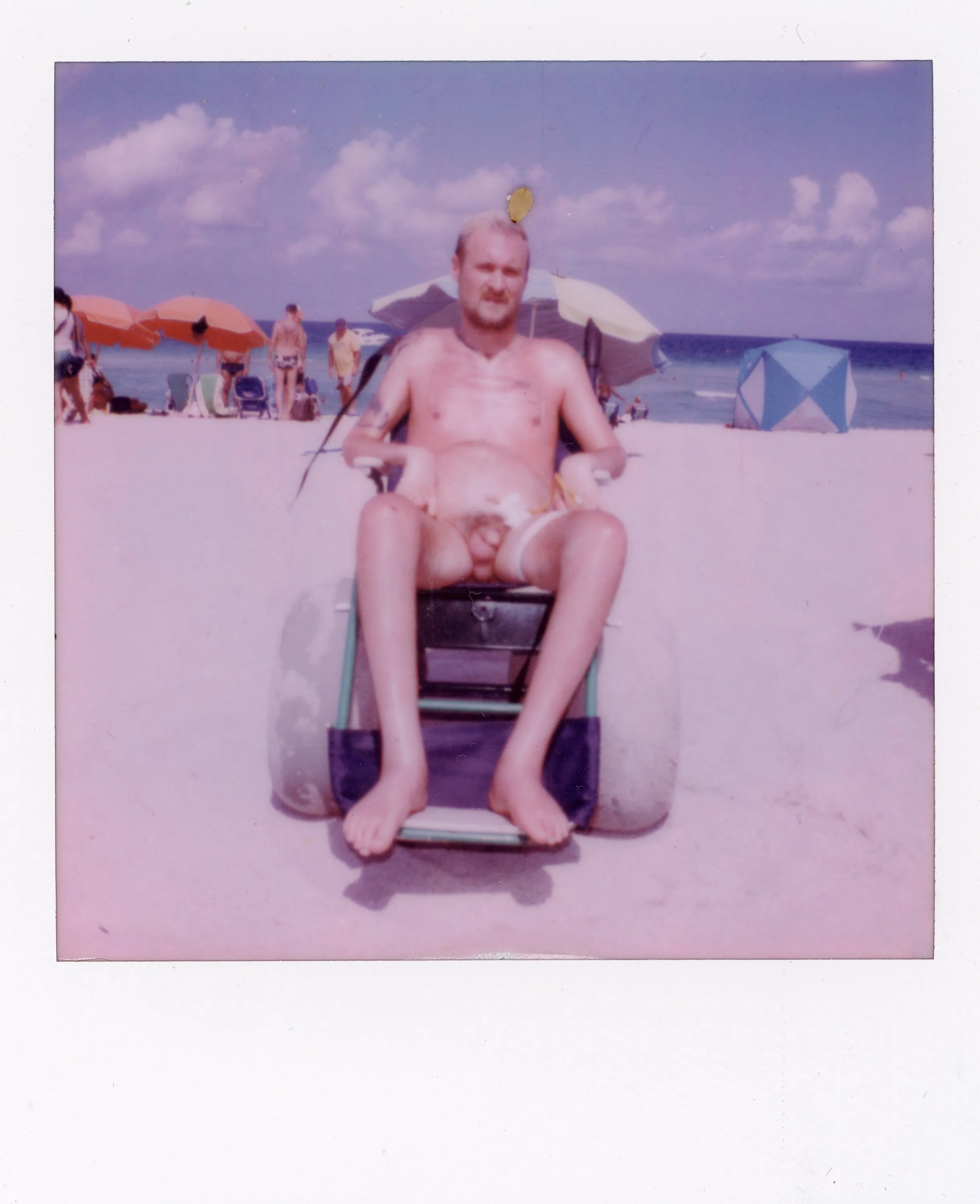 A man with a beard and no shirt sitting on a beach chair with a person with a tattoo. Beach umbrellas and other beachgoers are in the background on a sunny day.