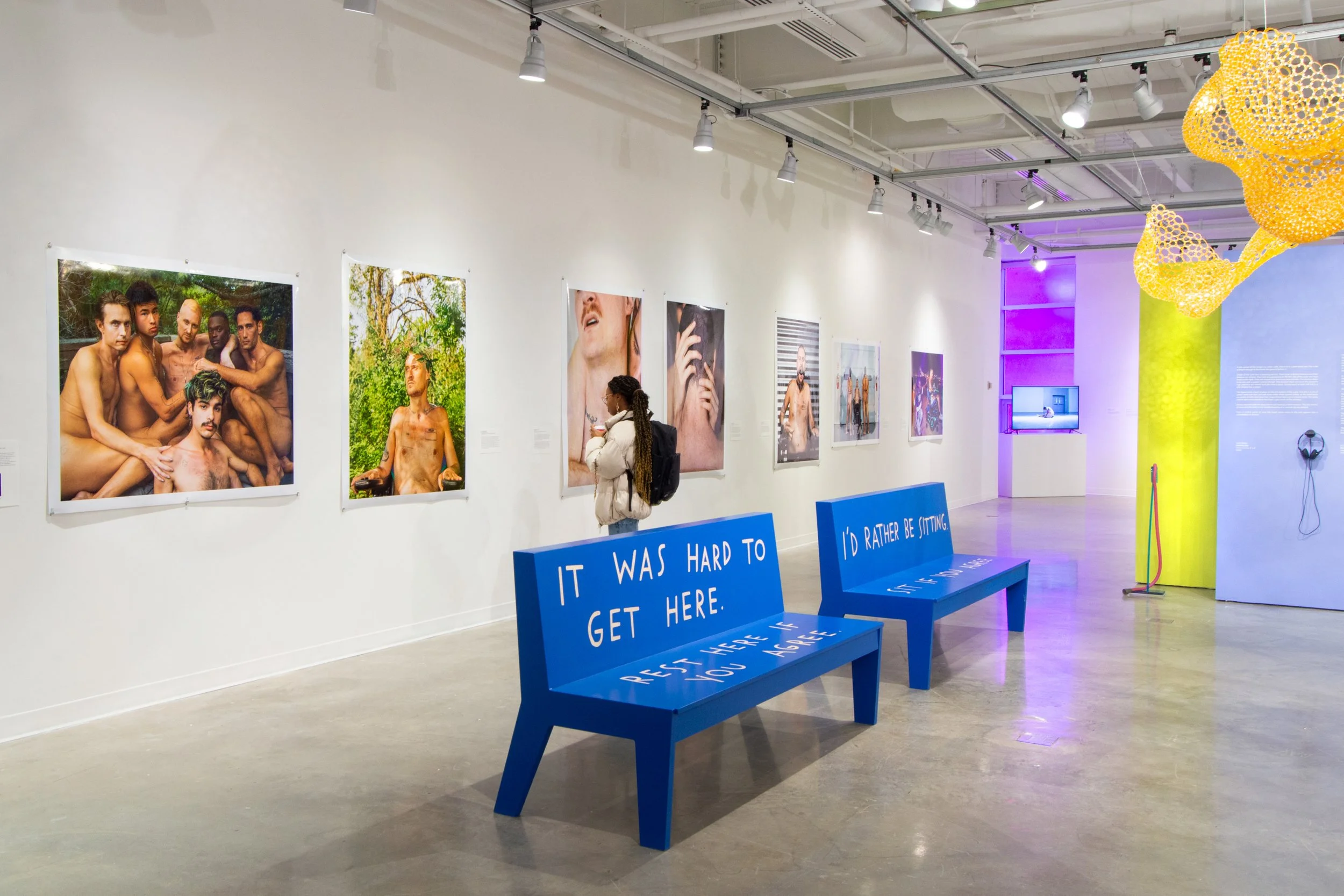 Gallery with photographs of shirtless men, contemporary art installations, and colorful seating with phrases, in a modern art exhibit space.