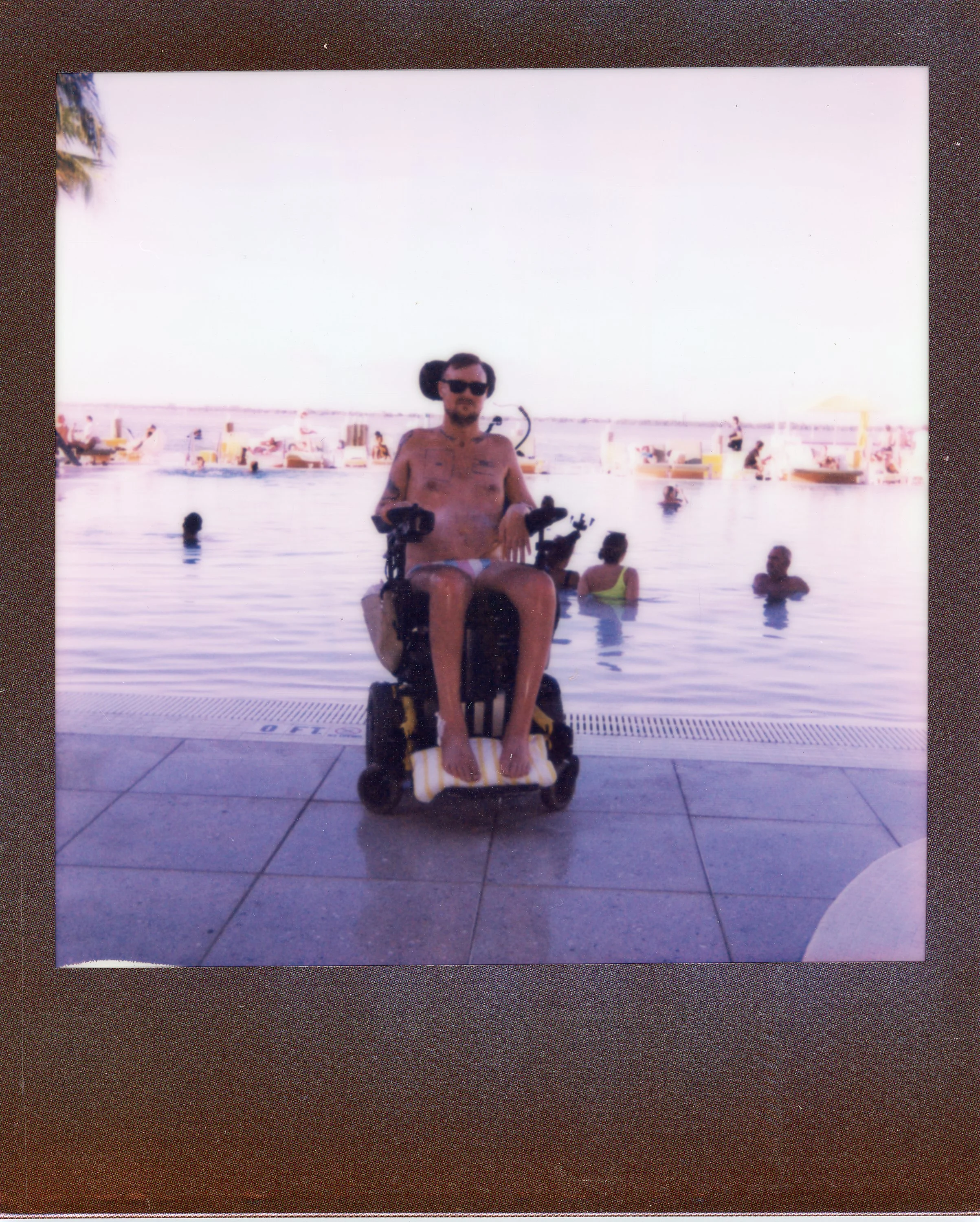 Man with a bicycle in a wheelchair at a poolside, wearing sunglasses and patterned swim trunks, with several people swimming and lounging in the pool and boats on the water in the background.