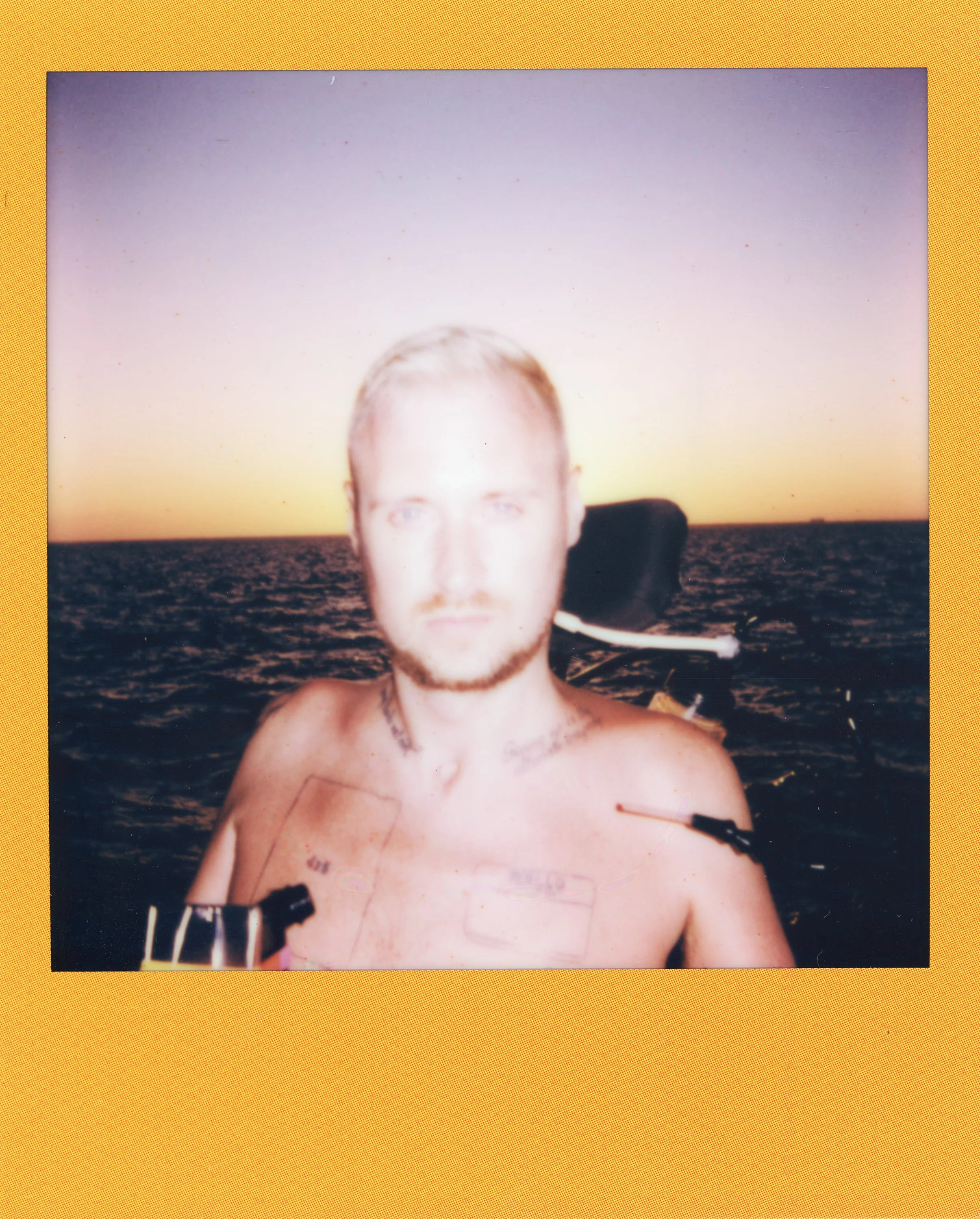 A young man with blond hair and a beard taking a selfie on a boat during sunset, with water and a distant horizon in the background.