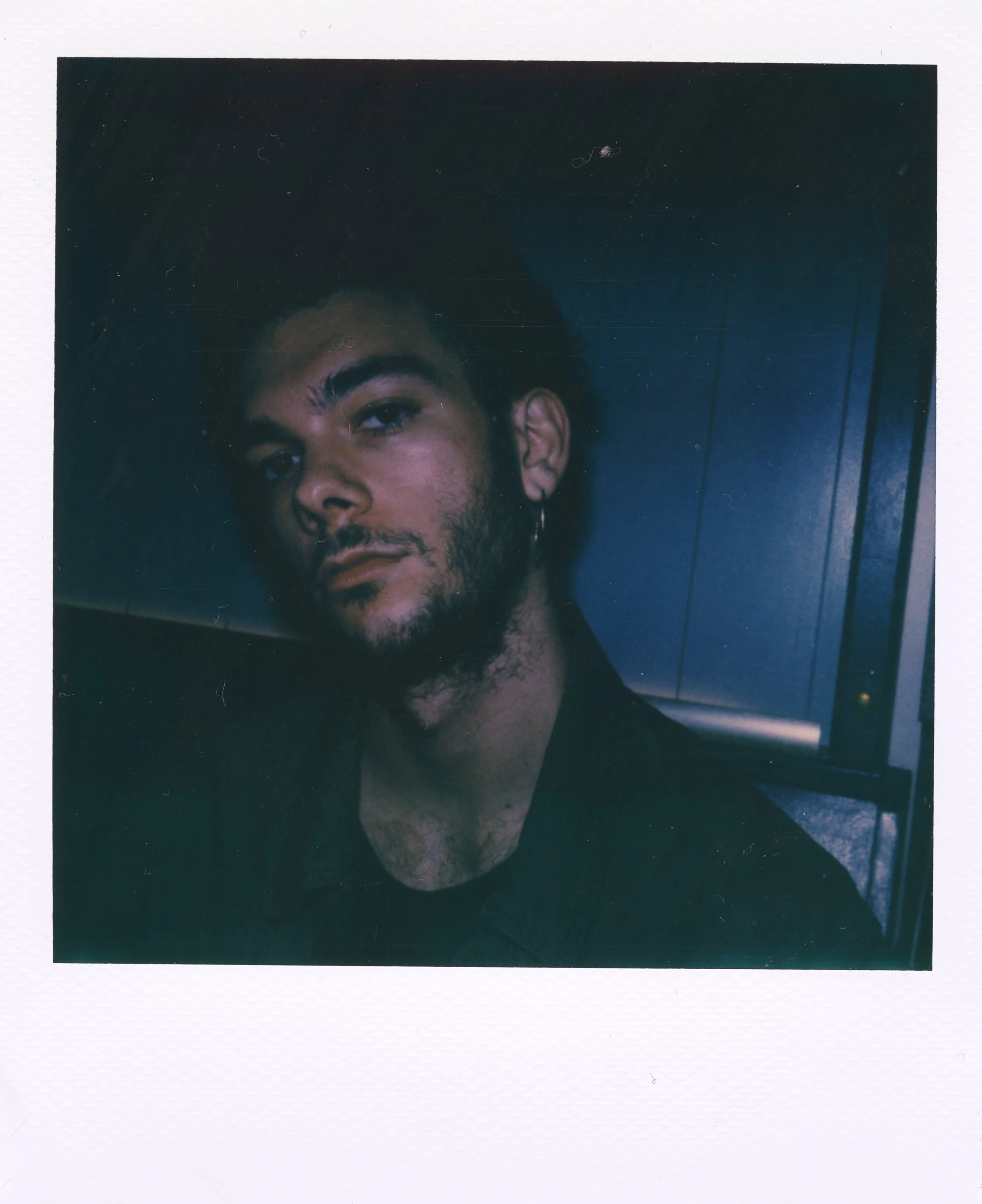 A young man with dark hair, a beard, and an earring in his left ear, looking at the camera with a serious expression, against a dark background.