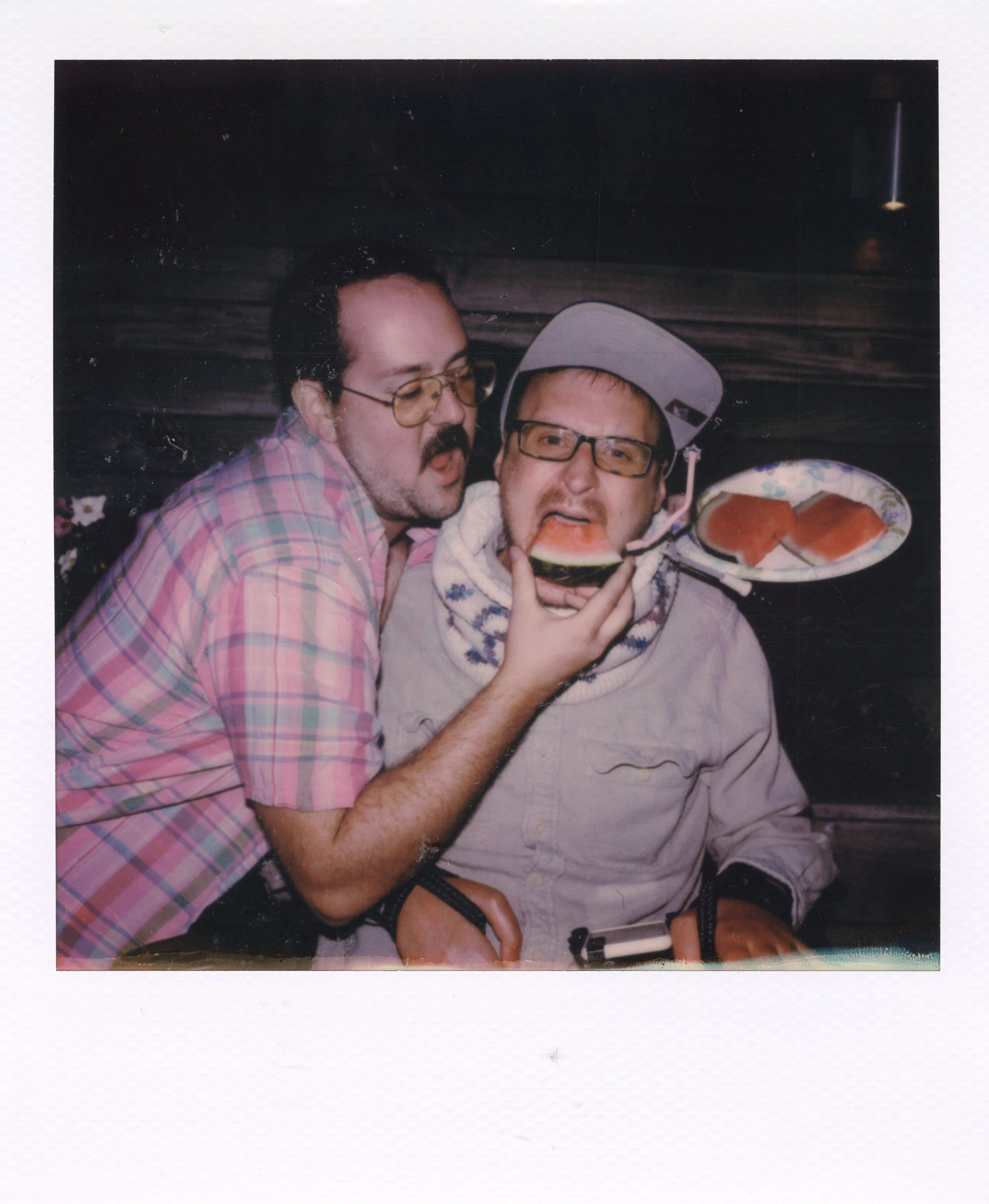 Two men with glasses engaging in a humorous moment, one holding a plate with food, the other pretending to eat watermelon from the plate.
