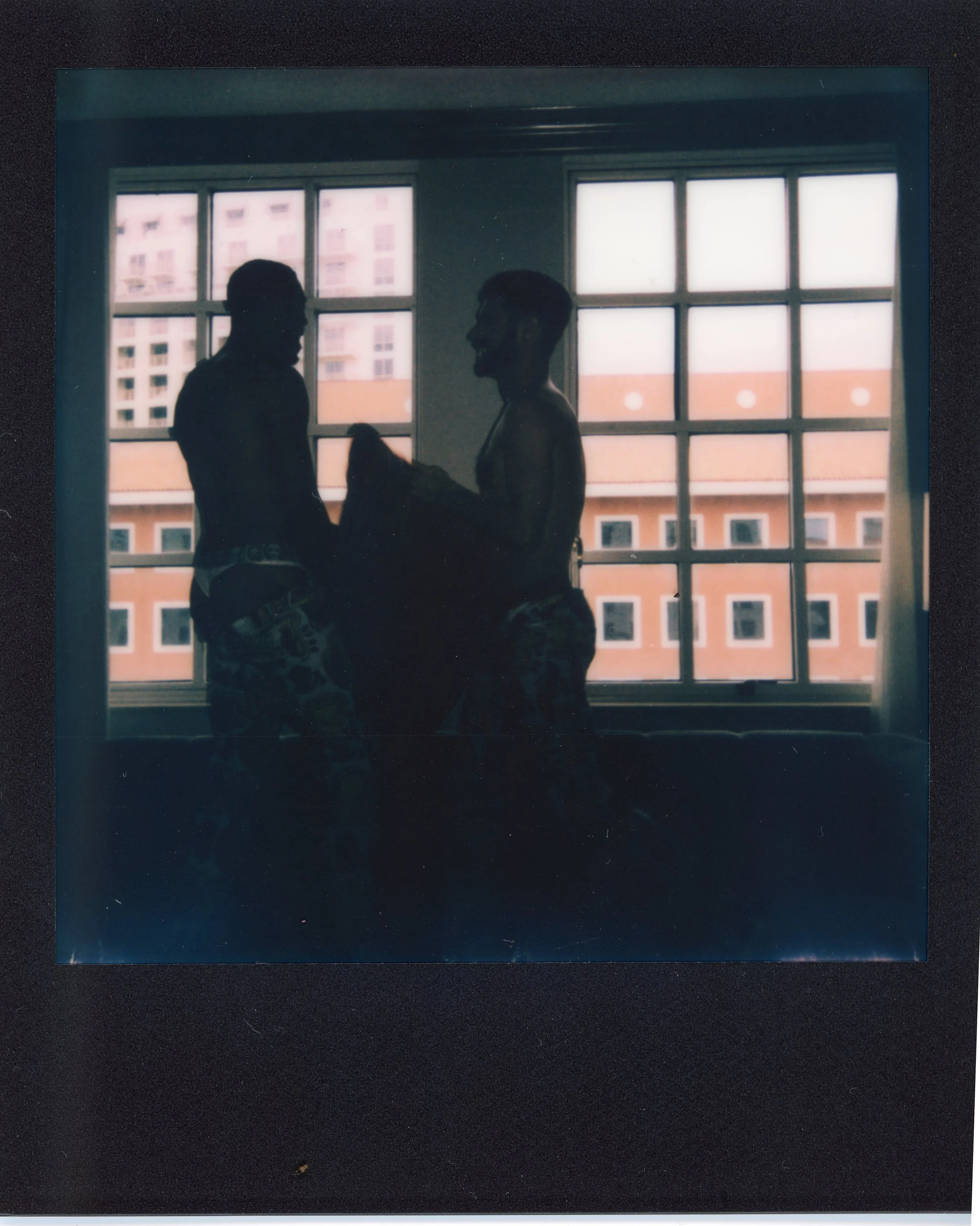 Silhouette of two shirtless men with underwear near a window, one holding an object, in a room with a cityscape background.