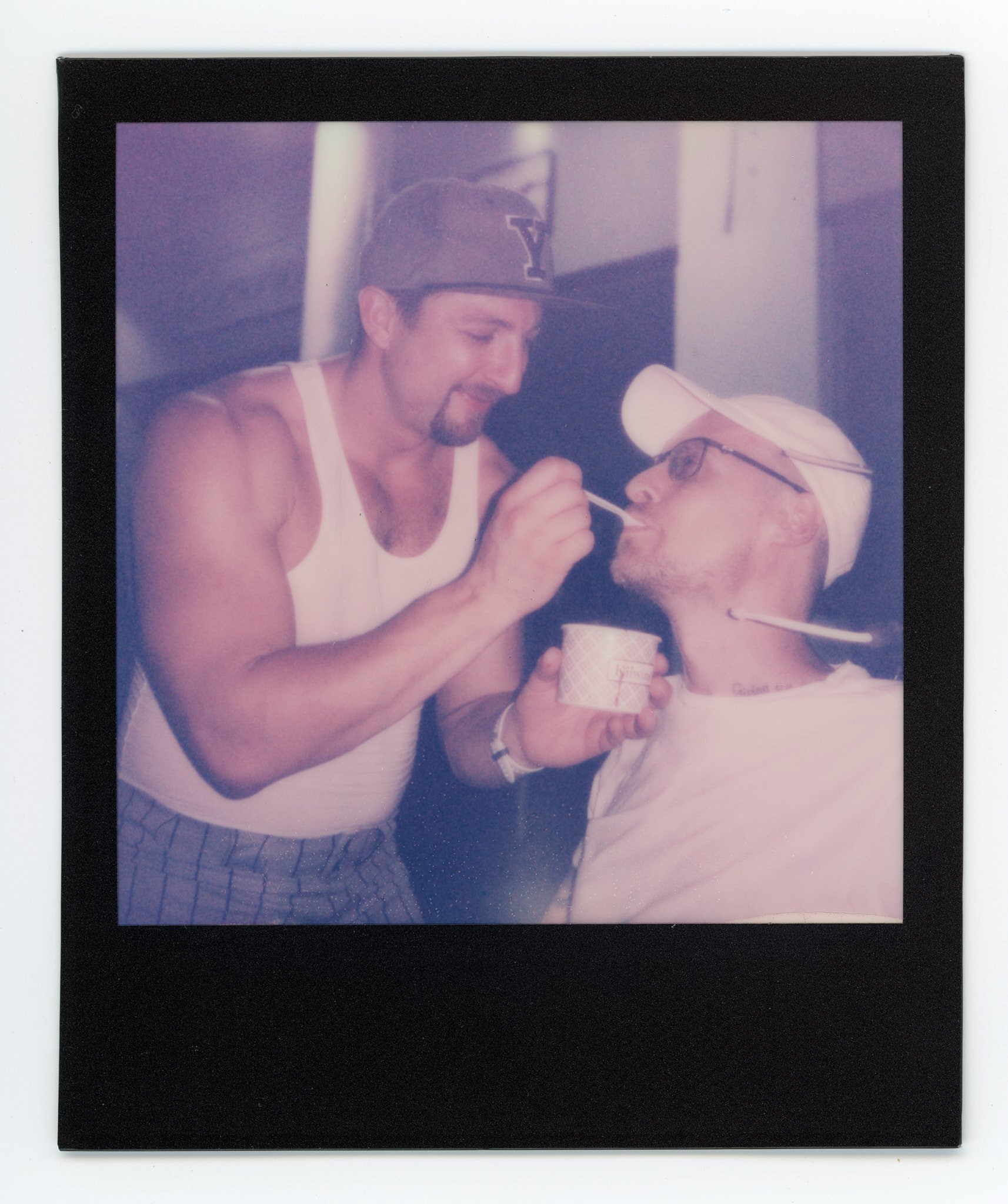 Two men, one wearing a Yale cap, are sharing a spoonful of ice cream. The man with the Yale cap is standing, while the other man, wearing glasses and a white cap, is seated.