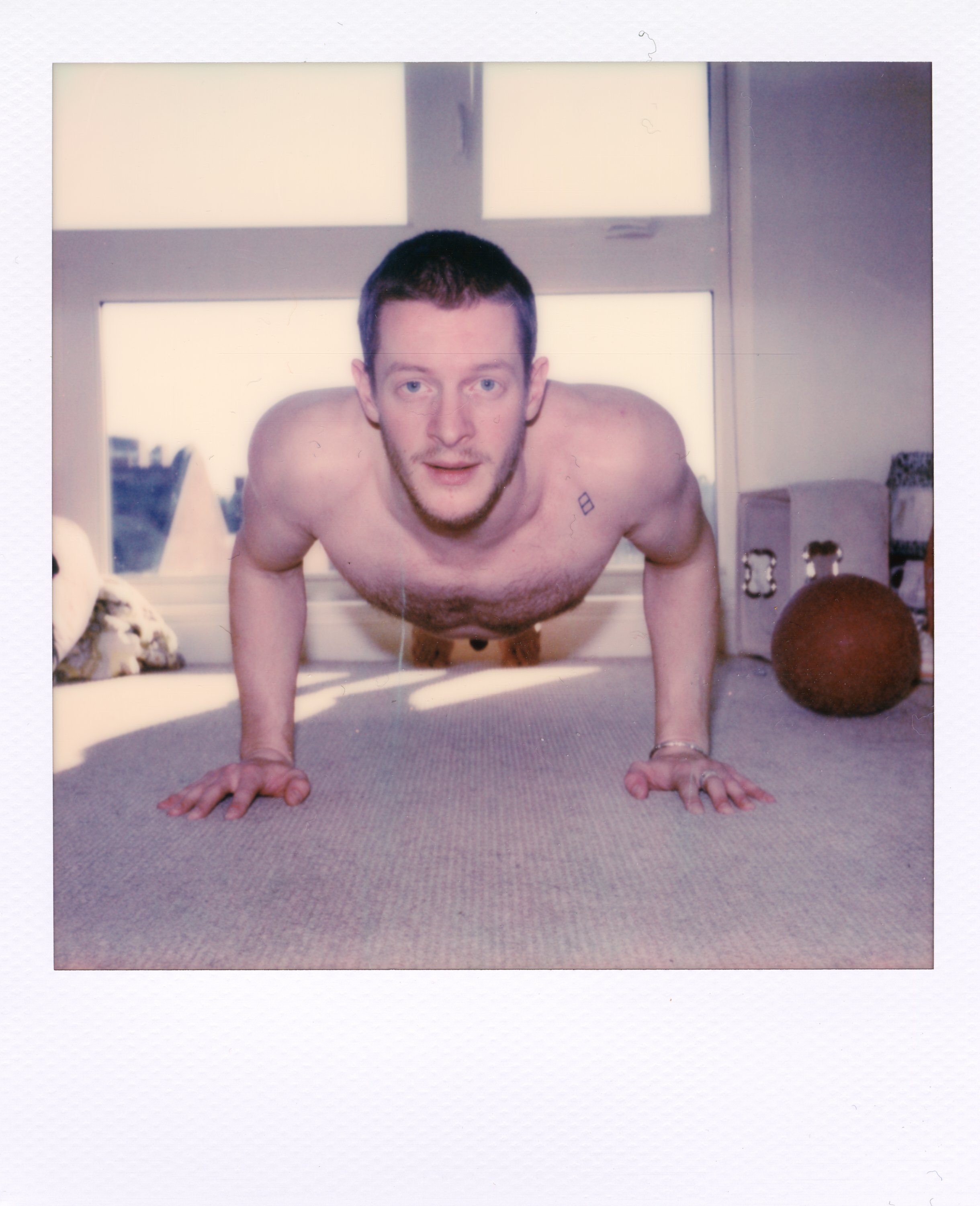A shirtless man with short dark hair and a beard doing push-ups on a carpeted floor in a room with large windows and various objects in the background.