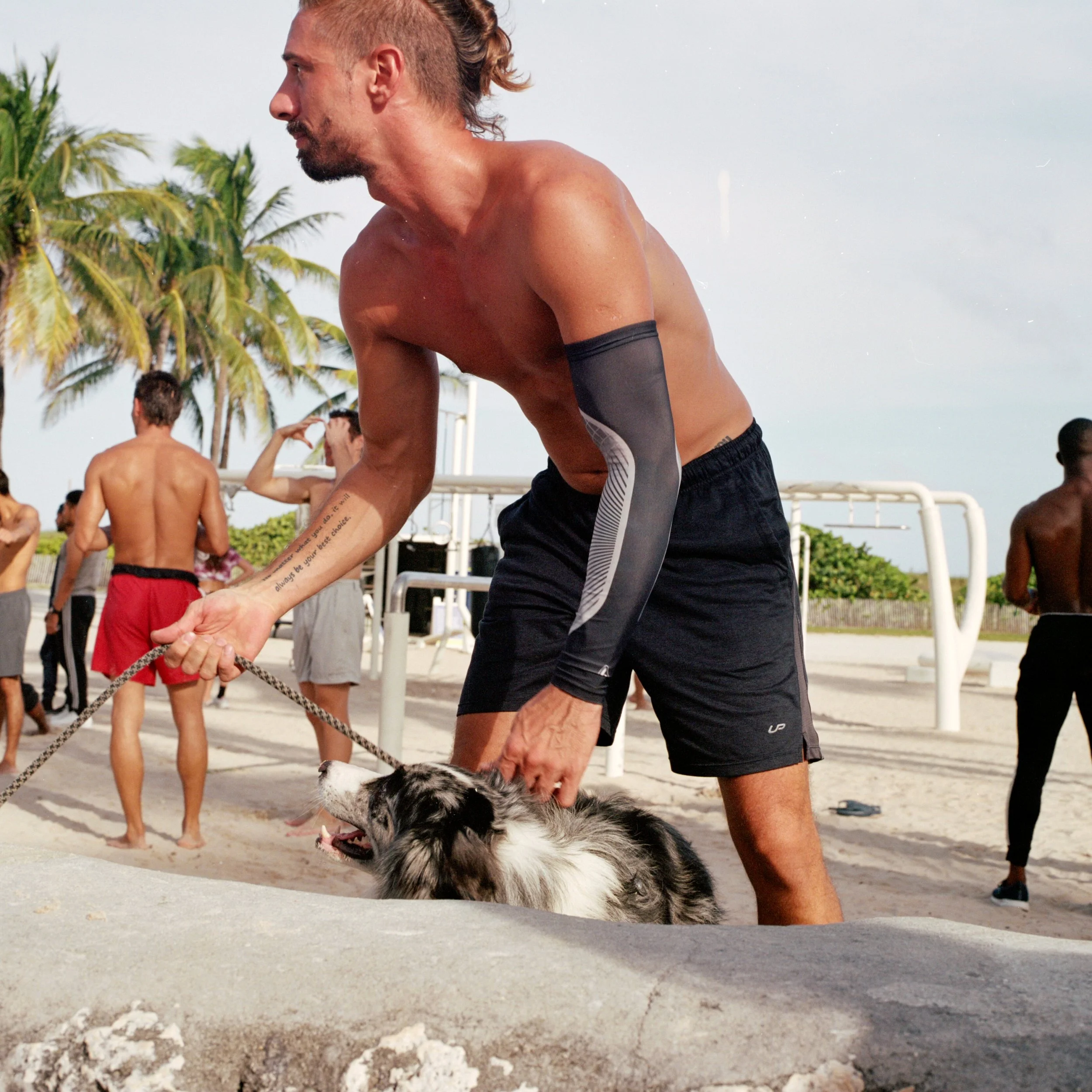 A shirtless man with tattoos on his arm holding a leash attached to a dog on a beach with several people in the background and palm trees.
