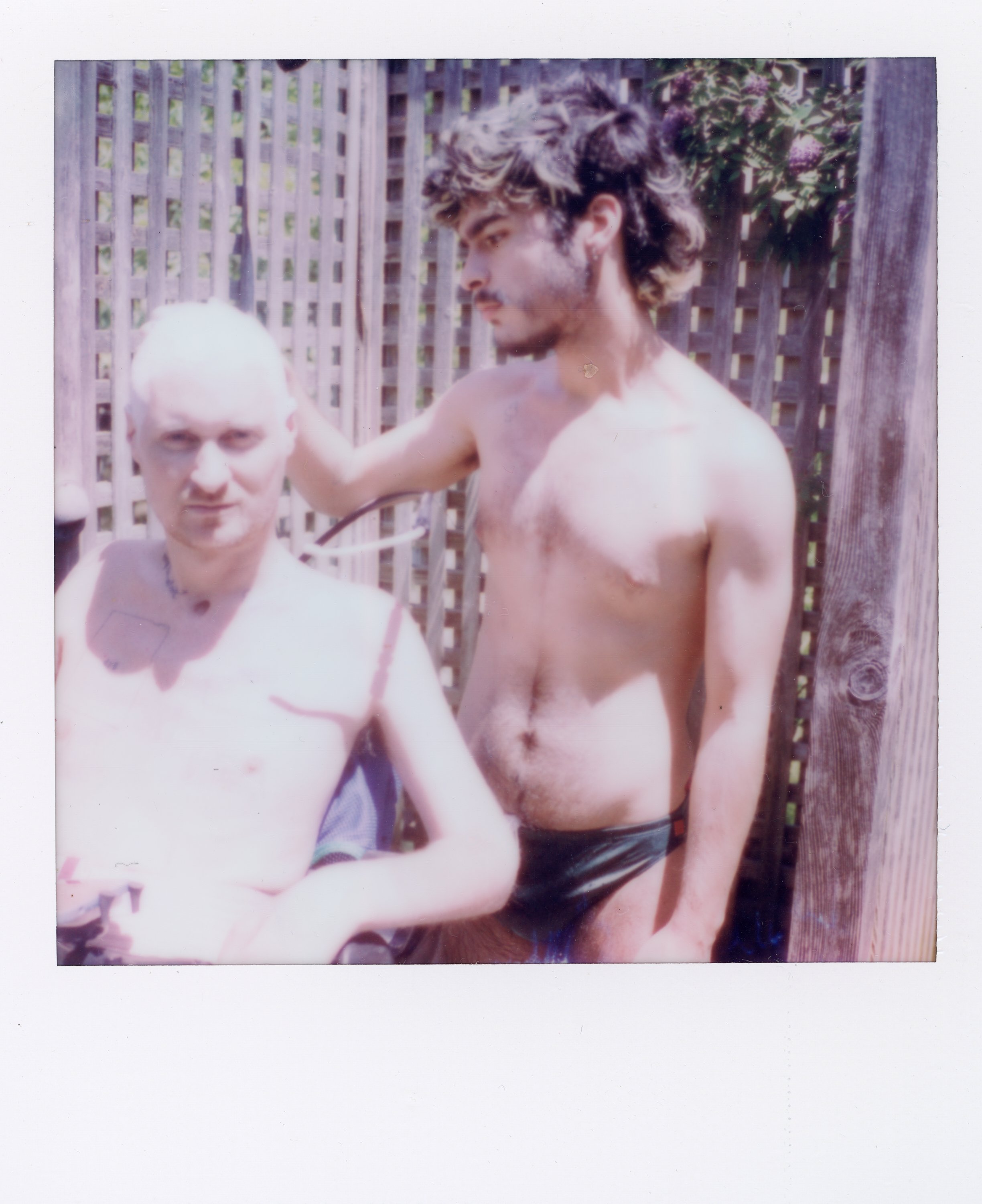 Two shirtless young men with one sitting and the other standing, in front of a wooden fence, outdoors on a sunny day.