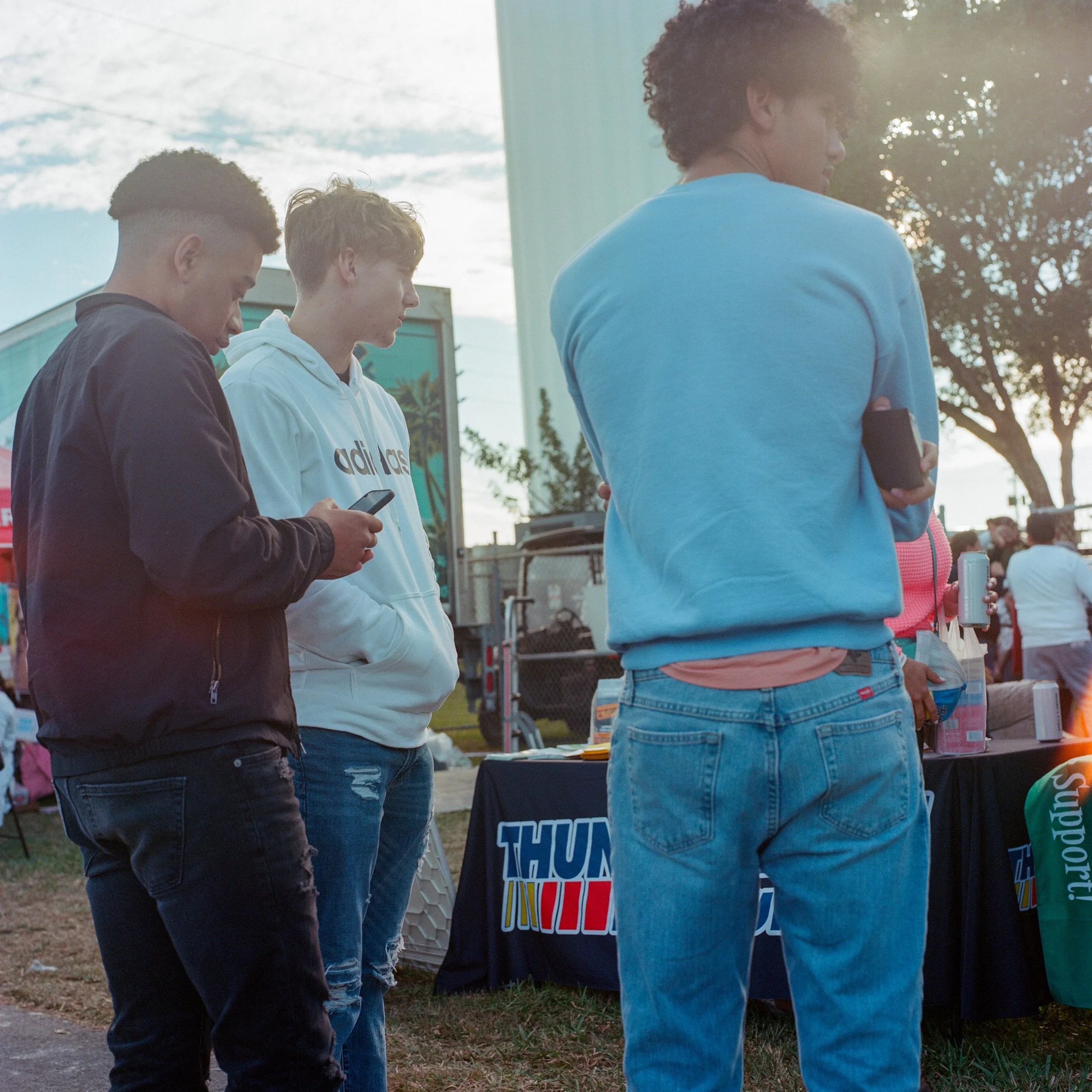 Three young men standing outdoors at a booth, two are looking at their phones and one has his arm crossed, with a crowd in the background.