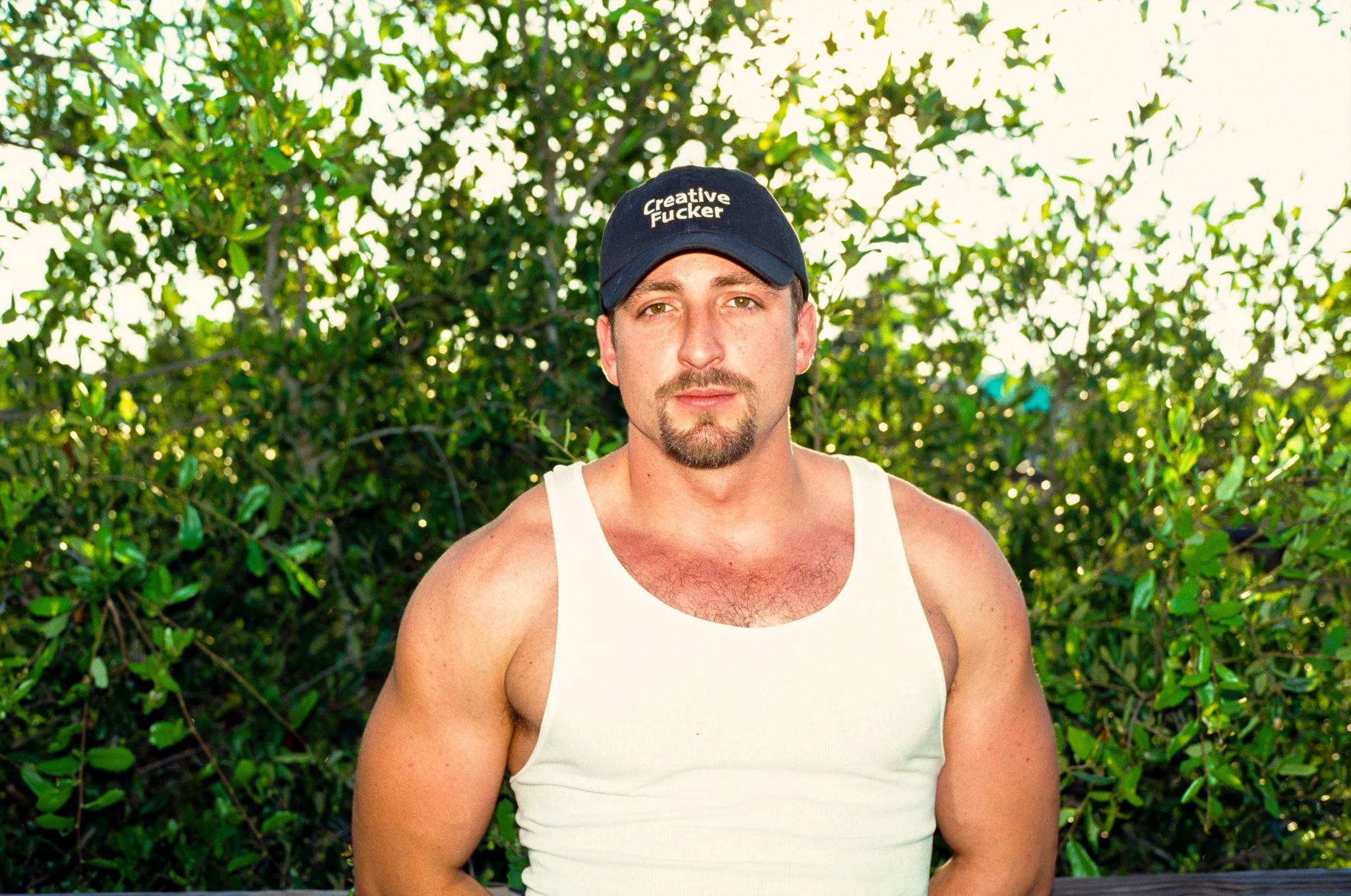 A man with a mustache and goatee wearing a white tank top and a black baseball cap with the words 'Creative Fcker' written on it, standing outdoors in front of green foliage.