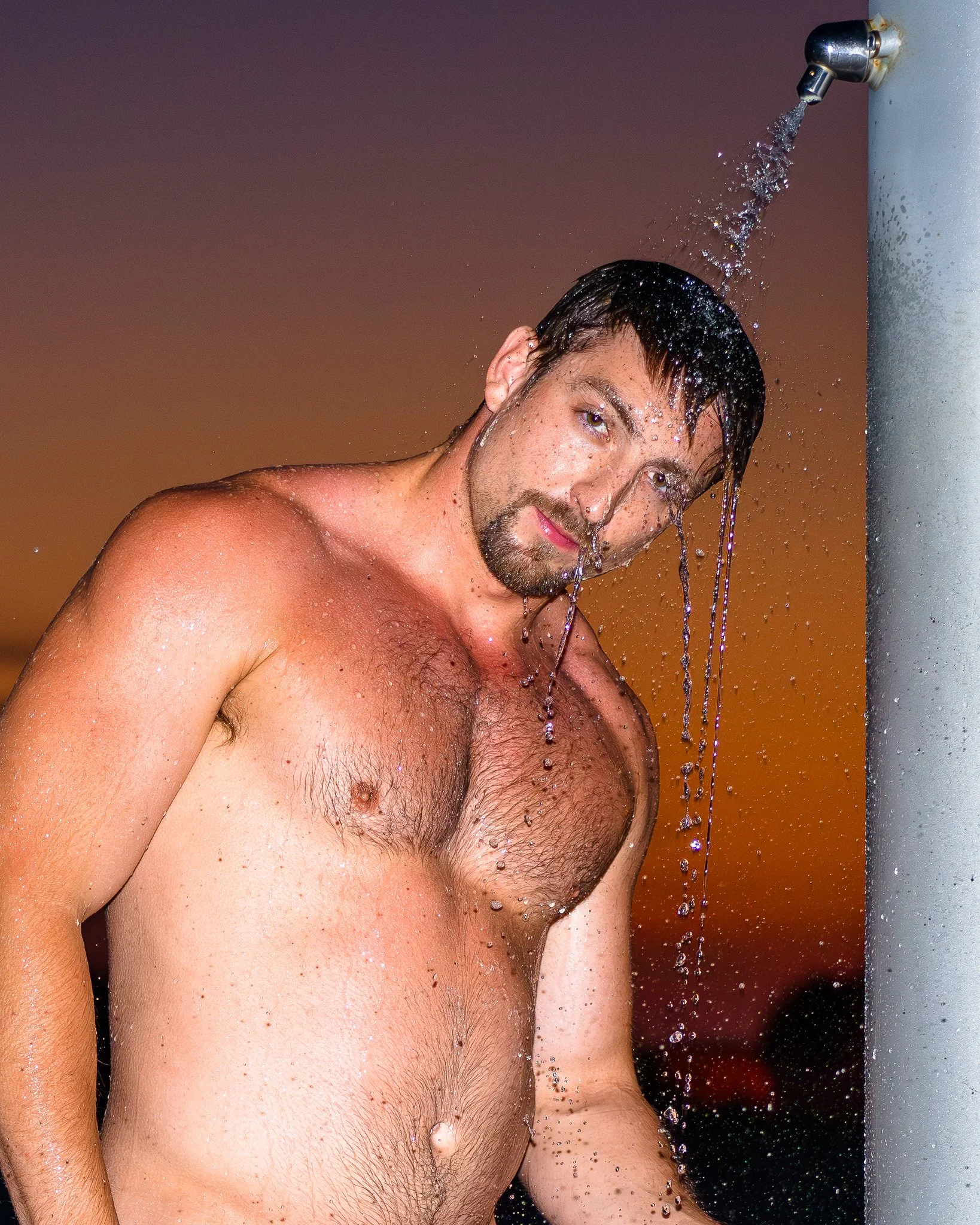 A shirtless man with dark hair and a beard takes an outdoor shower at sunset, with water running from the showerhead on his head.