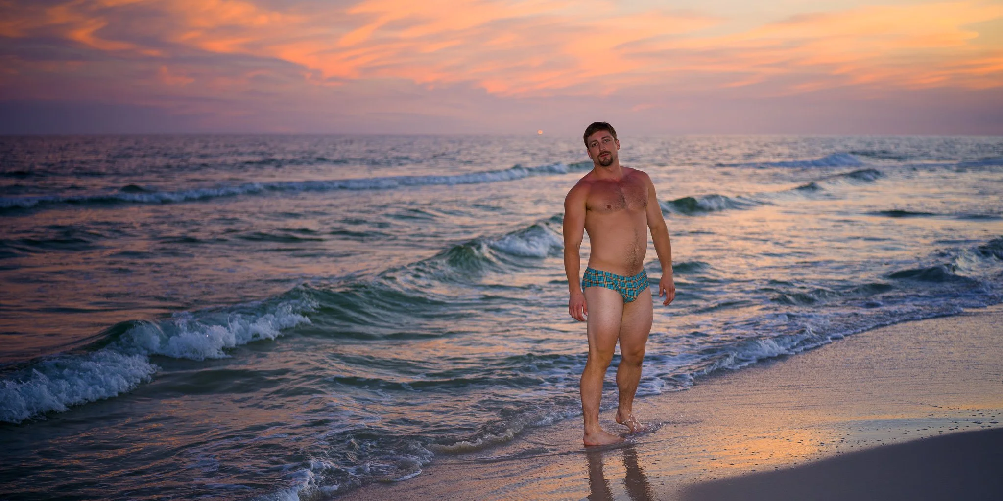 A man with a beard and short hair standing in shallow ocean water at sunset, wearing blue and green plaid swim trunks, with a pink and orange sky in the background.