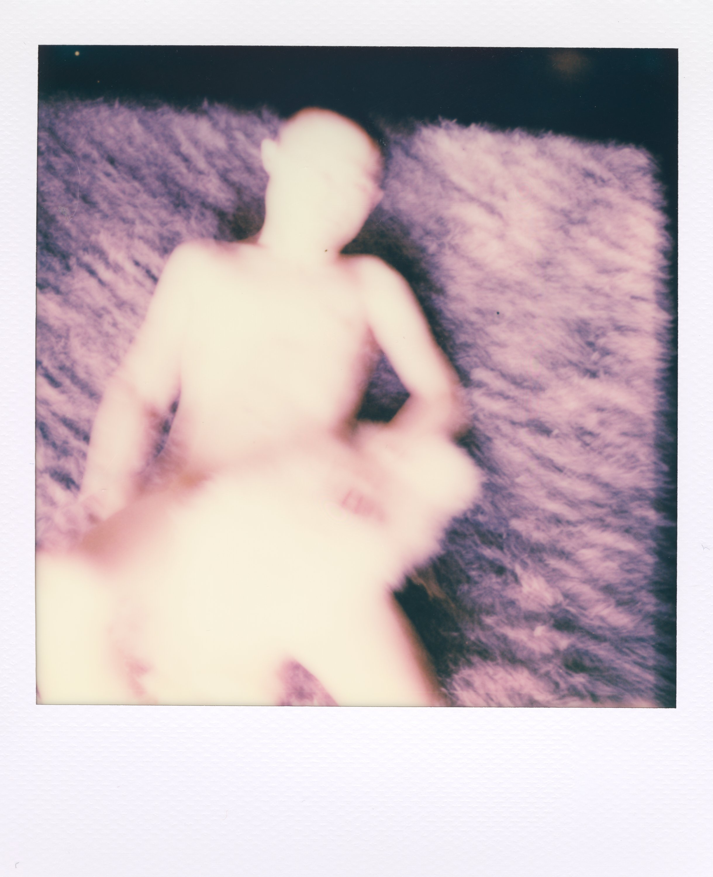 An intentionally blurred Polaroid photo of a person lying on a purple fluffy rug, with their body and face indistinct.