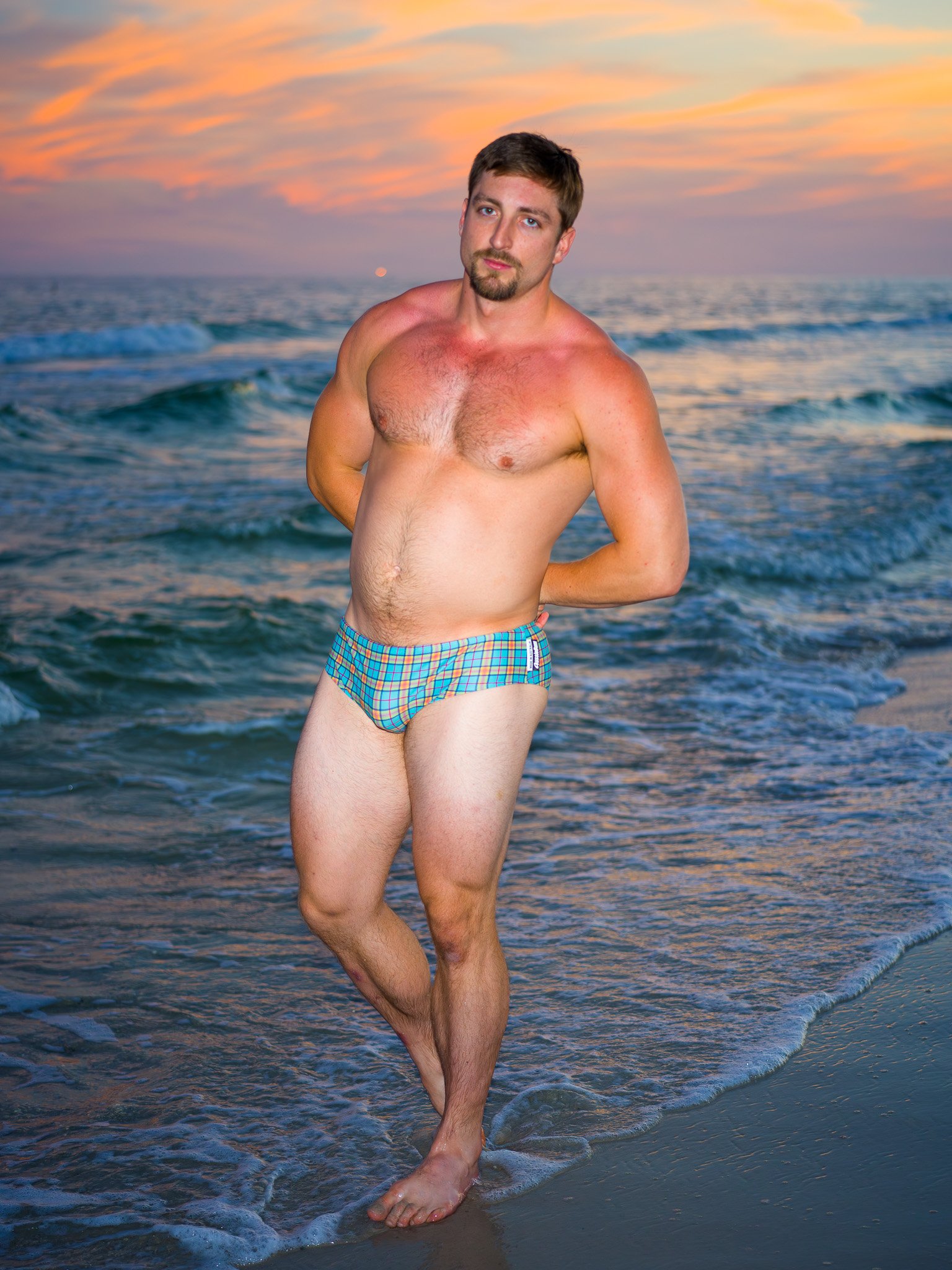 A man stands partially in the ocean water on a beach, wearing plaid swim trunks during a sunset or sunrise.