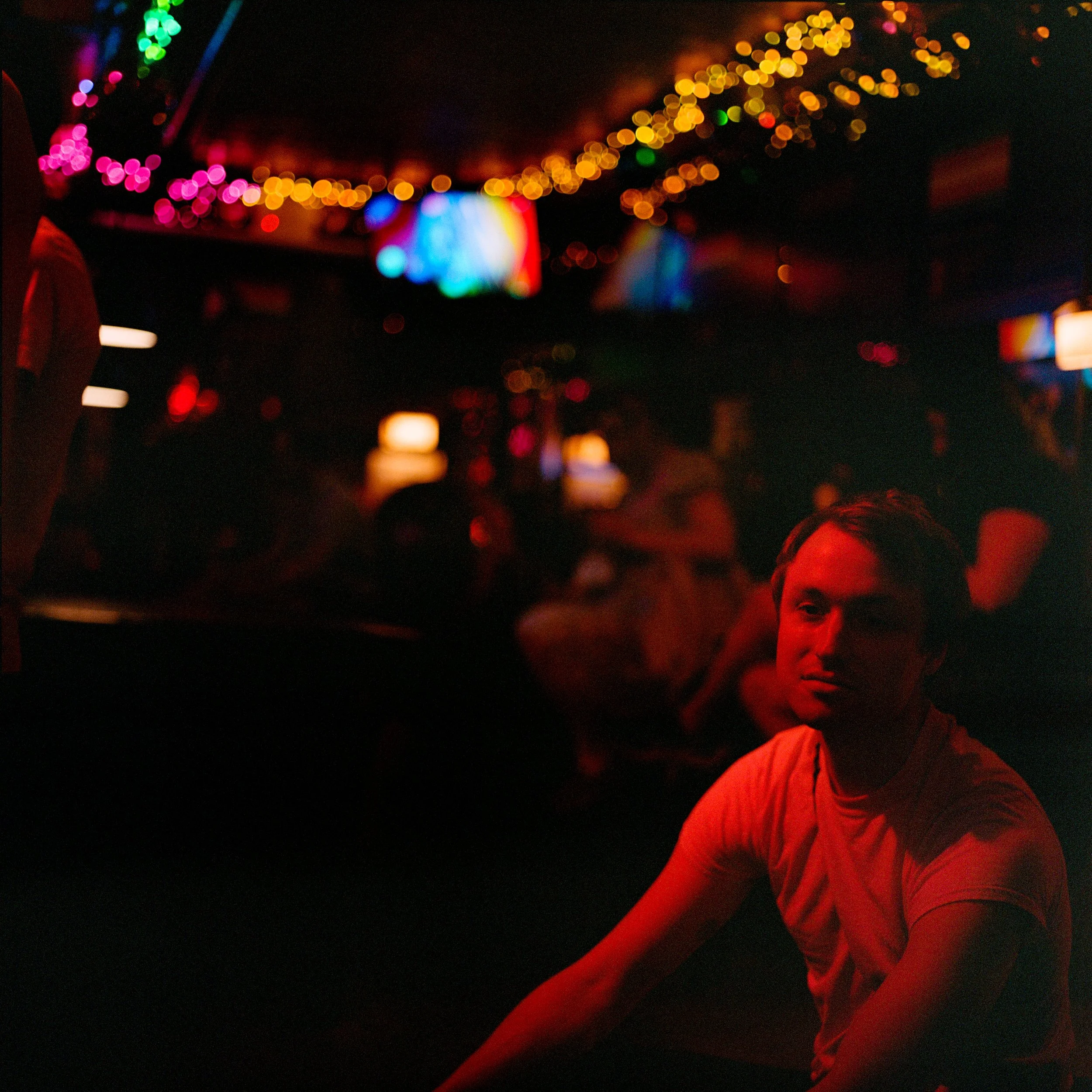 A young man with dark hair and a neutral expression sitting in a dimly lit bar or club with red lighting, with colorful blurred lights and people in the background.