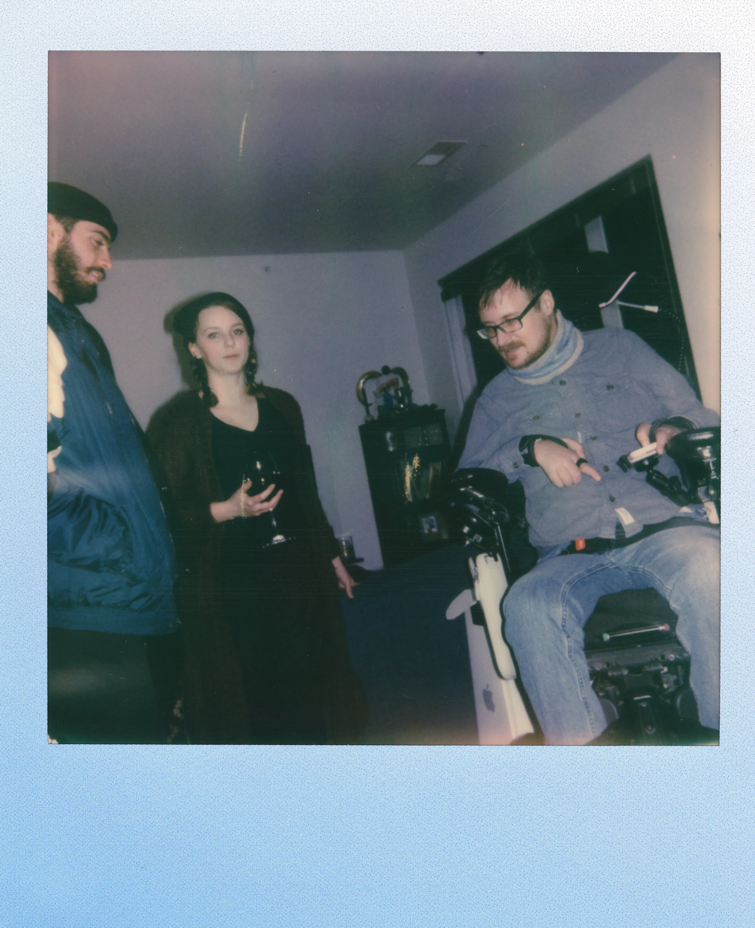 Three people standing around a man in a motorized wheelchair inside a home, with a man holding a smartphone and two women holding wine glasses.