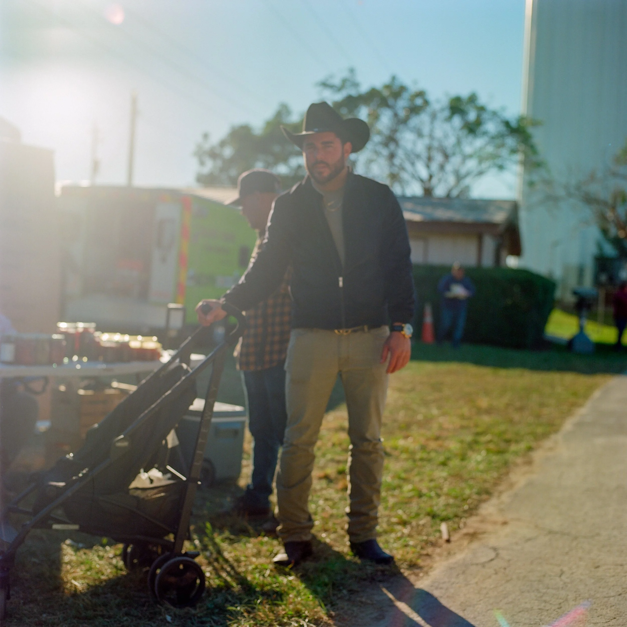 A man wearing a cowboy hat and casual clothing standing outdoors on a sunny day, with a stroller in front of him, and a vendor booth in the background.