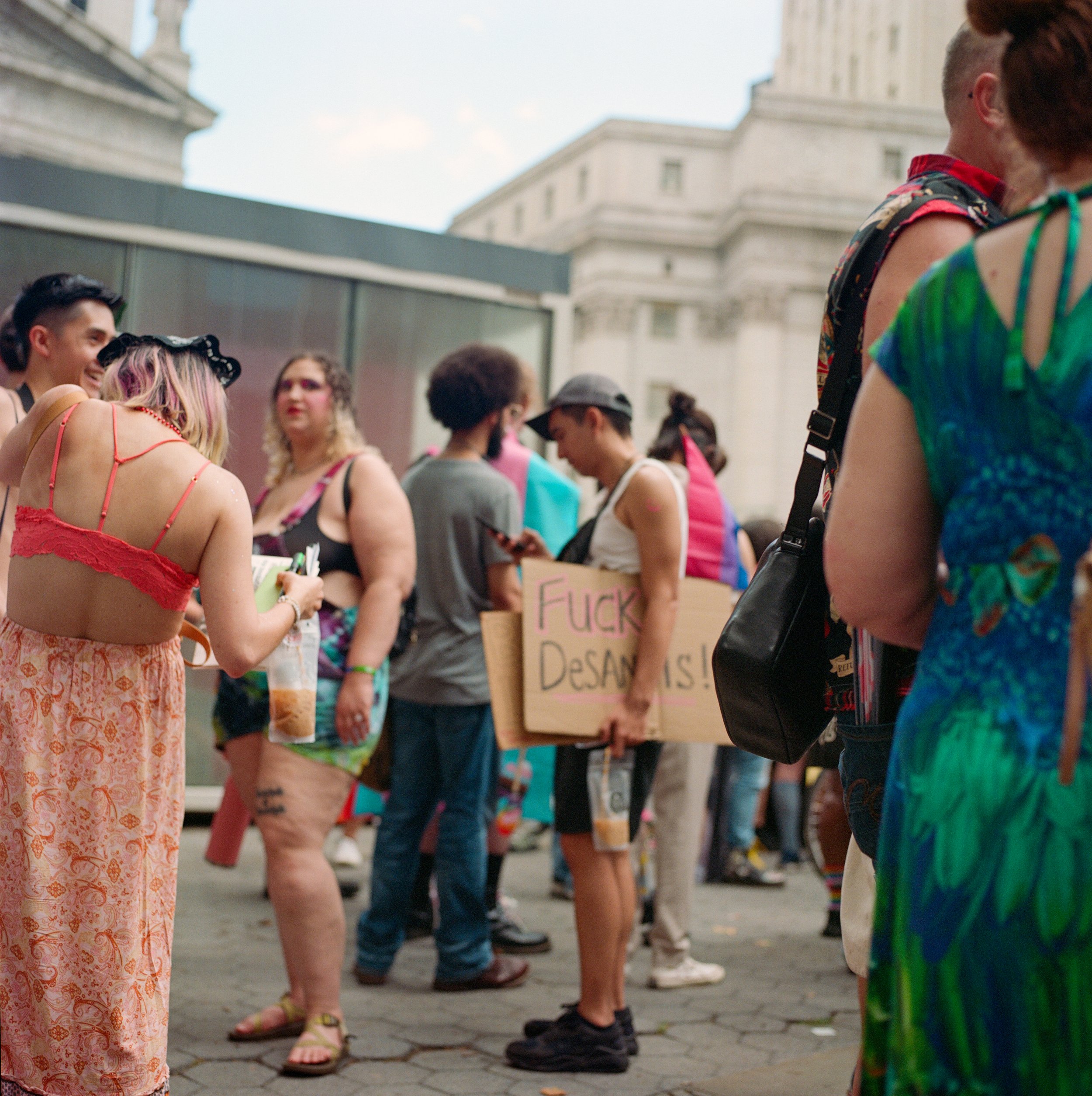 Group of people protesting outdoors, one person holding a sign with explicit language, some individuals engaged on their phones, in front of a historic building.
