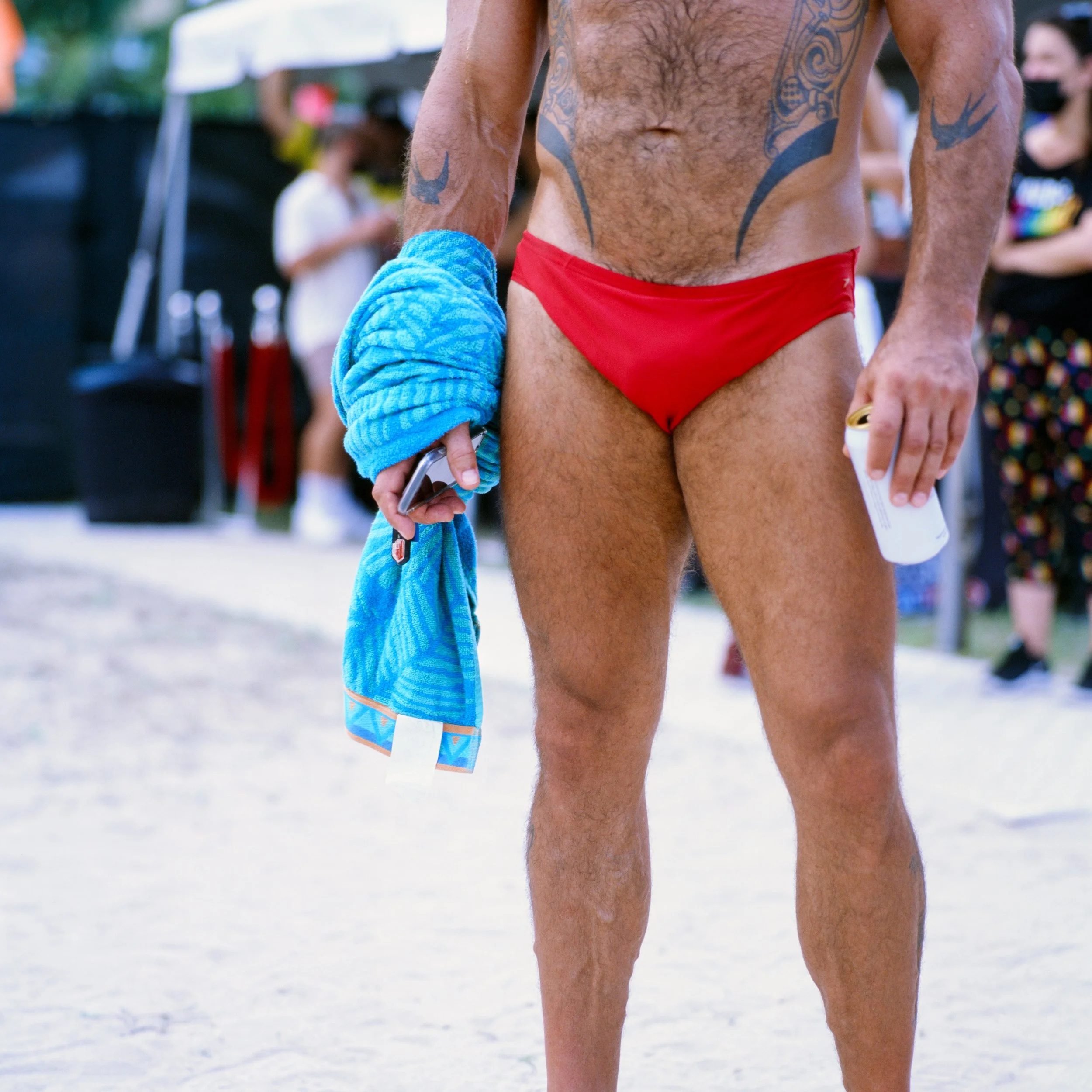 A man wearing red swim briefs holding a blue towel and a beverage can, standing on a sandy beach with other people in the background.